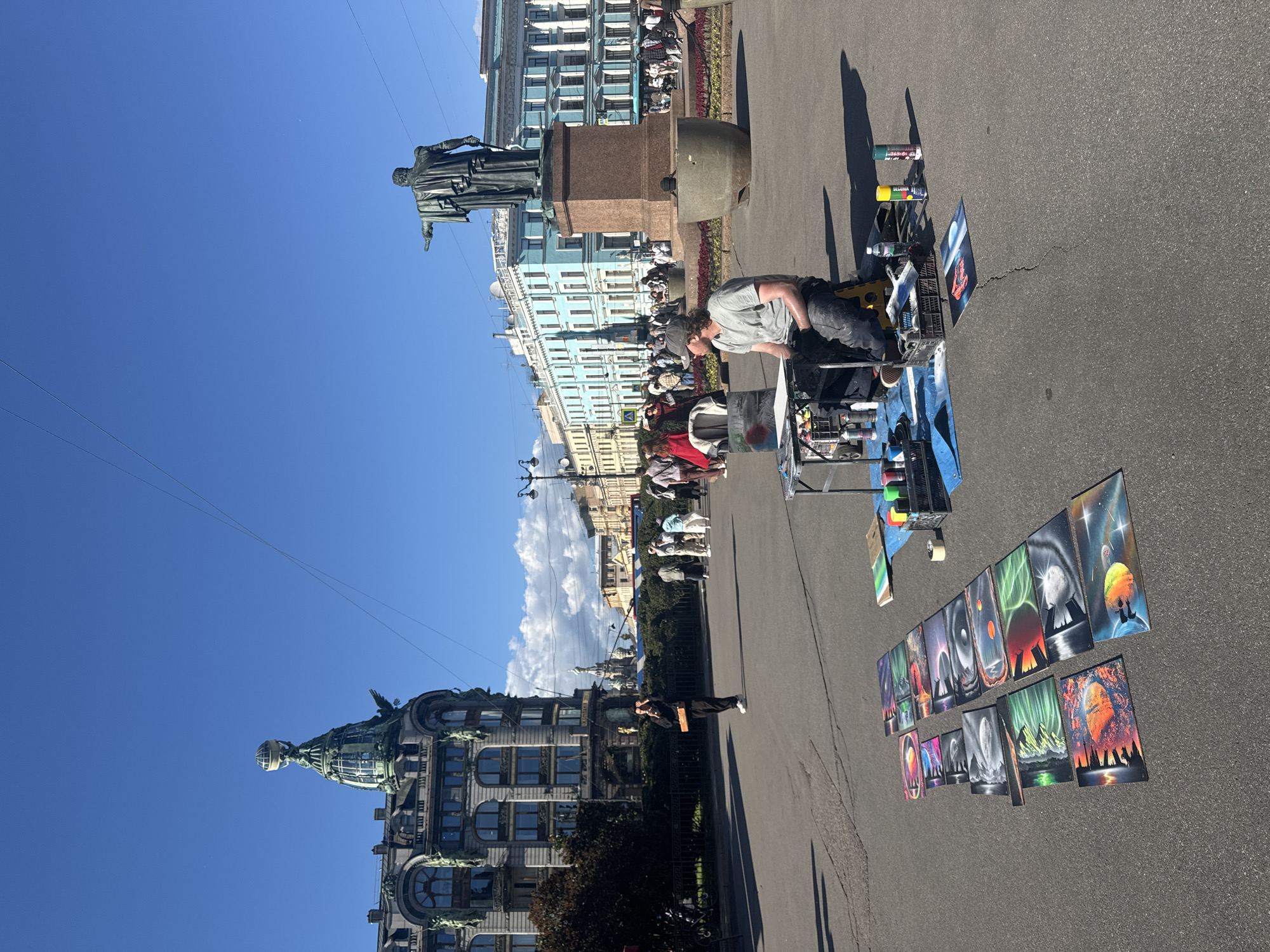 The image depicts an outdoor scene on a bright, sunny day in Saint Petersburg, Russia, likely on Palace Square. In the foreground, an artist is seated on a yellow stool behind a portable table, working on spray-painted artwork. Various finished spray-painted canvases are laid out on the asphalt in front of him, displaying colorful and detailed imagery, including landscapes and celestial scenes. Several cans of spray paint and other art supplies are arranged on the table and on the ground around him.
In the middle ground, a diverse group of pedestrians is walking through the square, some casually strolling, others appearing to be tourists. Behind the artist and the crowd, a tall bronze statue of a man in military attire stands on a stone pedestal, a prominent landmark in the square. Further in the background, several grand historic buildings with distinct architectural styles line the square. To the left, the iconic Singer House building with its distinctive glass globe and eagle sculpture is visible. The sky is a clear, vibrant blue, with a few wispy clouds. The scene conveys a lively atmosphere of a public space in a major city, with the artist adding a creative element to the urban landscape.