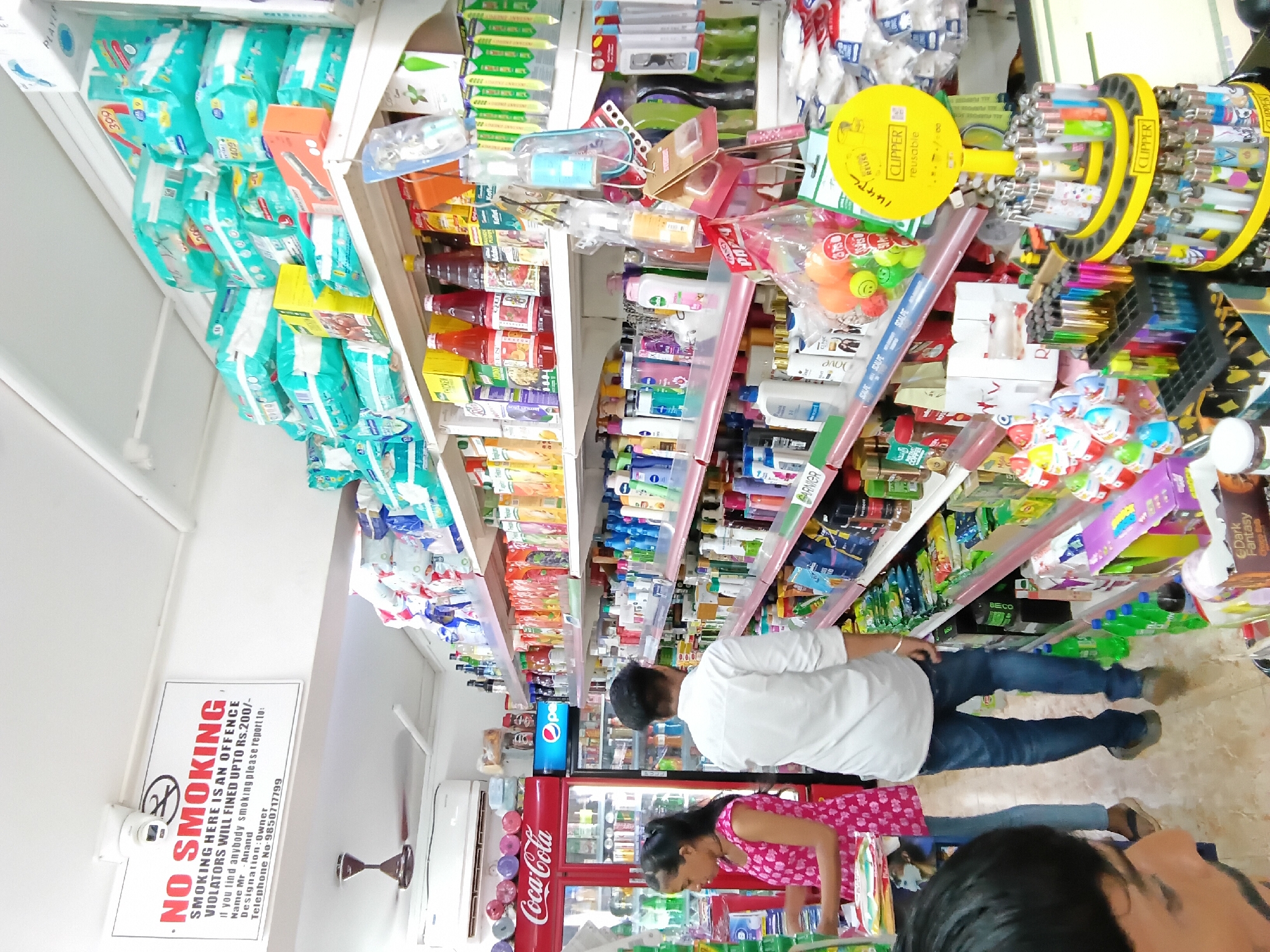 The image shows the interior of a store in Oxel, India. Aisles are lined with shelves stocked with a variety of products, including diapers, beverages (Coca-Cola), snacks, and personal care items. A man in a white shirt and jeans stands in one of the aisles. A woman in a pink floral dress is bending over. A "No Smoking" sign is visible on the wall. A display of Clipper lighters is present.