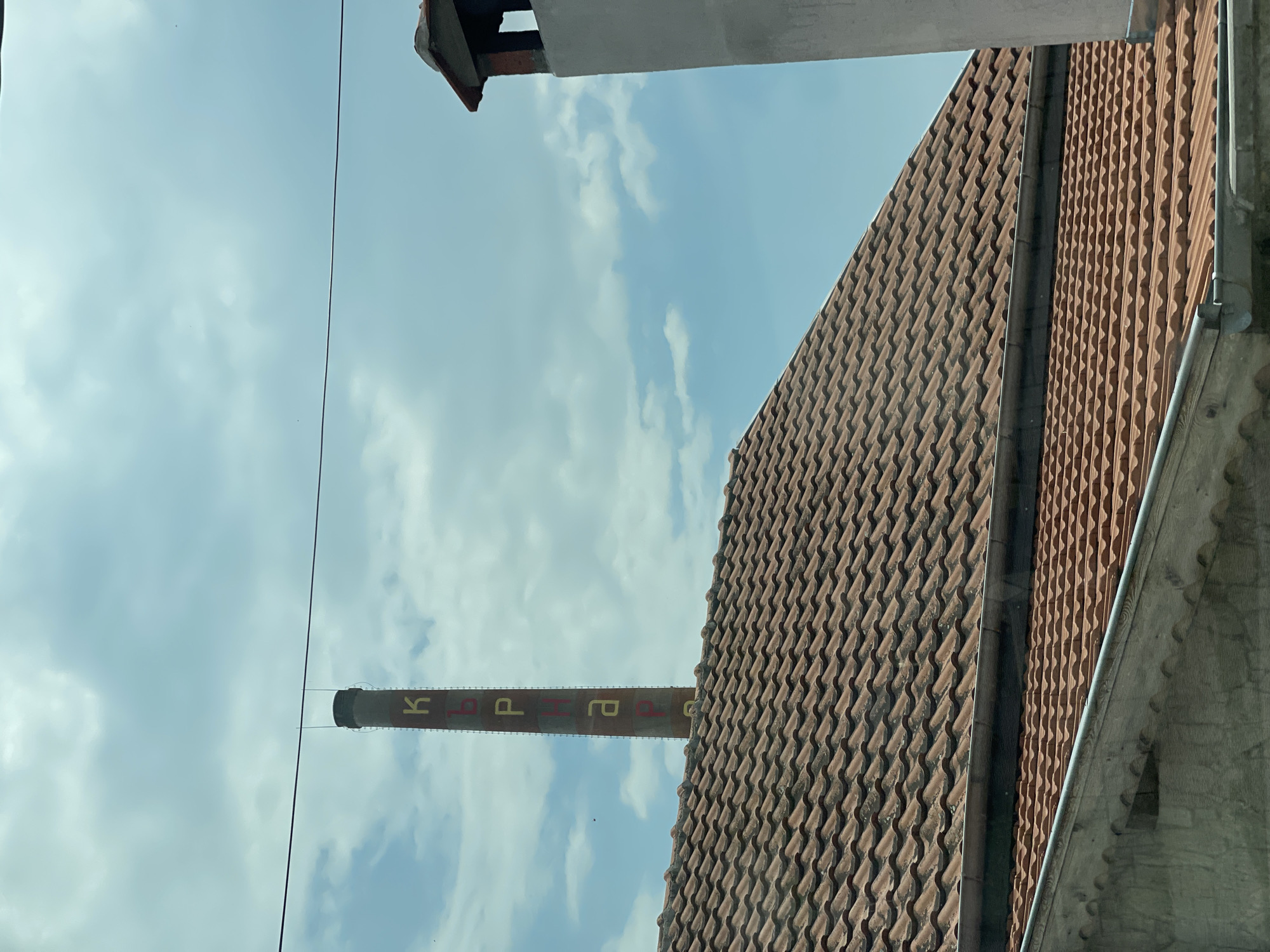 The image captures an outdoor daytime scene dominated by an industrial chimney and multiple rooftops against a cloudy sky. A tall, slender, reddish-brown brick chimney stands centrally, featuring vertically stacked Cyrillic letters painted in alternating yellow and red: 'К', 'Ъ', 'Р', 'Н', 'А', 'Р', 'Е', which spell "КЪРНАРЕ" (Karnare). This identifies the location as Karnare, Bulgaria. Below the chimney, several layers of terracotta-tiled rooftops with visible gutters are present, indicating residential or other buildings. On the far right, a partial view of a light grey, rectangular chimney or vent structure with a darker opening and a reddish cap is visible. A single dark utility wire extends horizontally across the upper portion of the image, positioned in front of the industrial chimney. The sky above is partly cloudy with patches of blue. No people or specific actions are depicted.