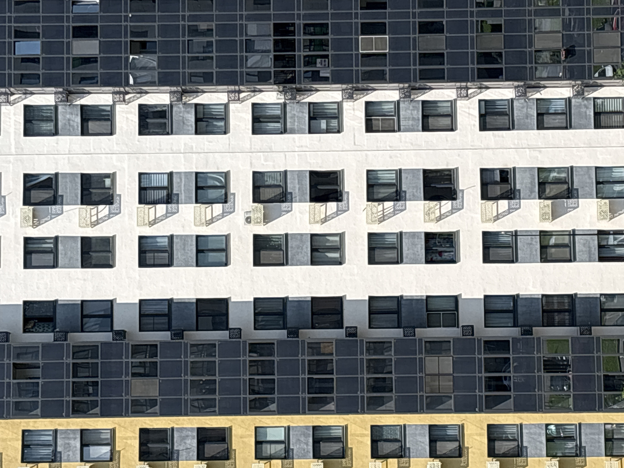 This is a close-up, high-angle view of a modern, multi-story residential building, likely located in Kazan, Russia. The building features a repeating pattern of windows and balconies across its facade. The facade is divided vertically into distinct sections with different cladding: a section of yellow material on the left, a dark gray, grid-like section in the center-left, a predominantly white section in the center, and a dark gray, glass-covered section on the right.
The windows are uniformly sized and arranged in vertical stacks. Many of them are dark, suggesting they are closed or have tinted glass, reflecting the surrounding environment. Some windows show glimpses of interior blinds or curtains. Several balconies, some with visible railing or glass enclosures, are also present. Small, cream-colored air conditioning units are mounted on the exterior walls beneath some of the windows. The building is bathed in sunlight, casting distinct shadows from the balconies and window frames onto the facade, highlighting the architectural details. The overall impression is of a densely built urban environment.