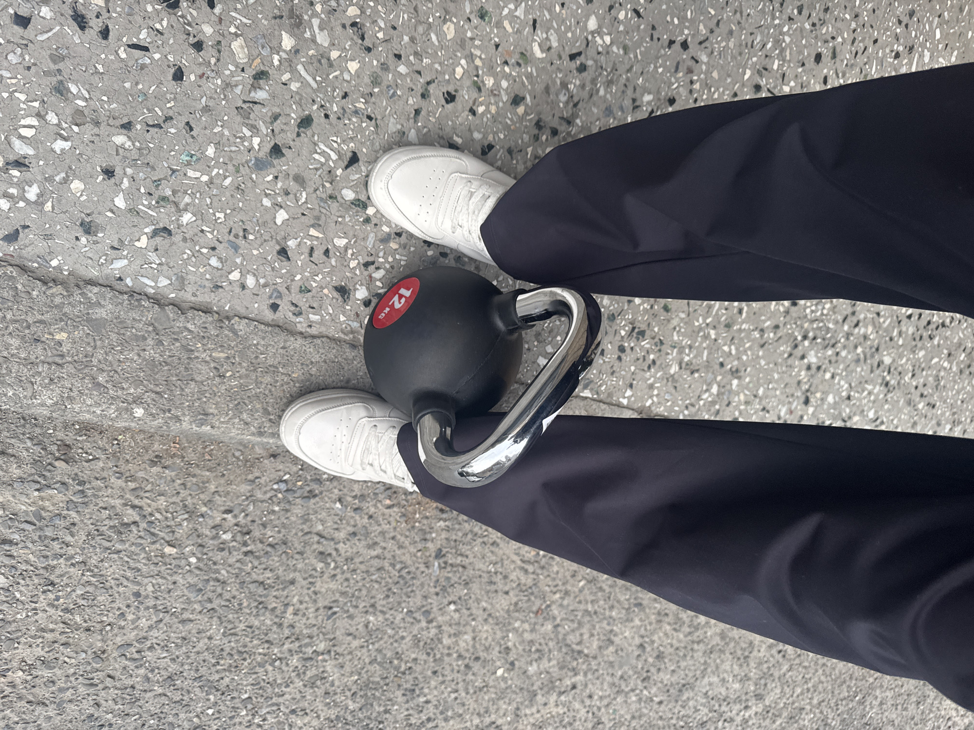 An overhead shot captures the lower body of a person standing outdoors, likely in an urban setting in Tbilisi, Georgia. The individual wears dark blue trousers and clean white sneakers. A black kettlebell with a shiny chrome handle rests between their feet. A red circular label on the kettlebell is clearly visible, indicating its weight as "12 KG." The ground is a composite of two types of concrete pavement. The predominant surface is a light grey terrazzo-like concrete embedded with various sizes of white, grey, and dark aggregate. A distinct seam or crack separates this from a darker, coarser-grained concrete area in the foreground, suggesting a change in paving or a curb. The lighting is bright and even, characteristic of daytime conditions, with no harsh shadows. The person appears to be standing statically, with no specific activity in progress.