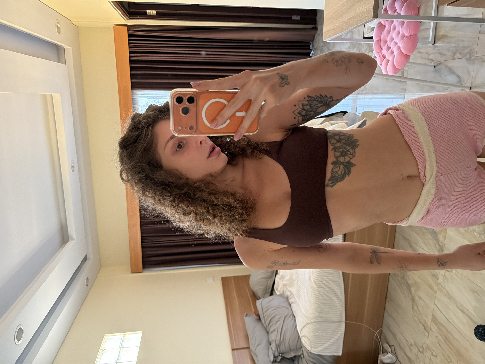 A woman is taking a selfie in a bedroom. She is wearing a brown sports bra and pink shorts. She has curly hair and tattoos on her arms and torso. The bedroom has a bed with gray bedding, brown curtains, and a white ceiling. A pink seat cushion is visible. The location is possibly in North Kuta, Indonesia.
