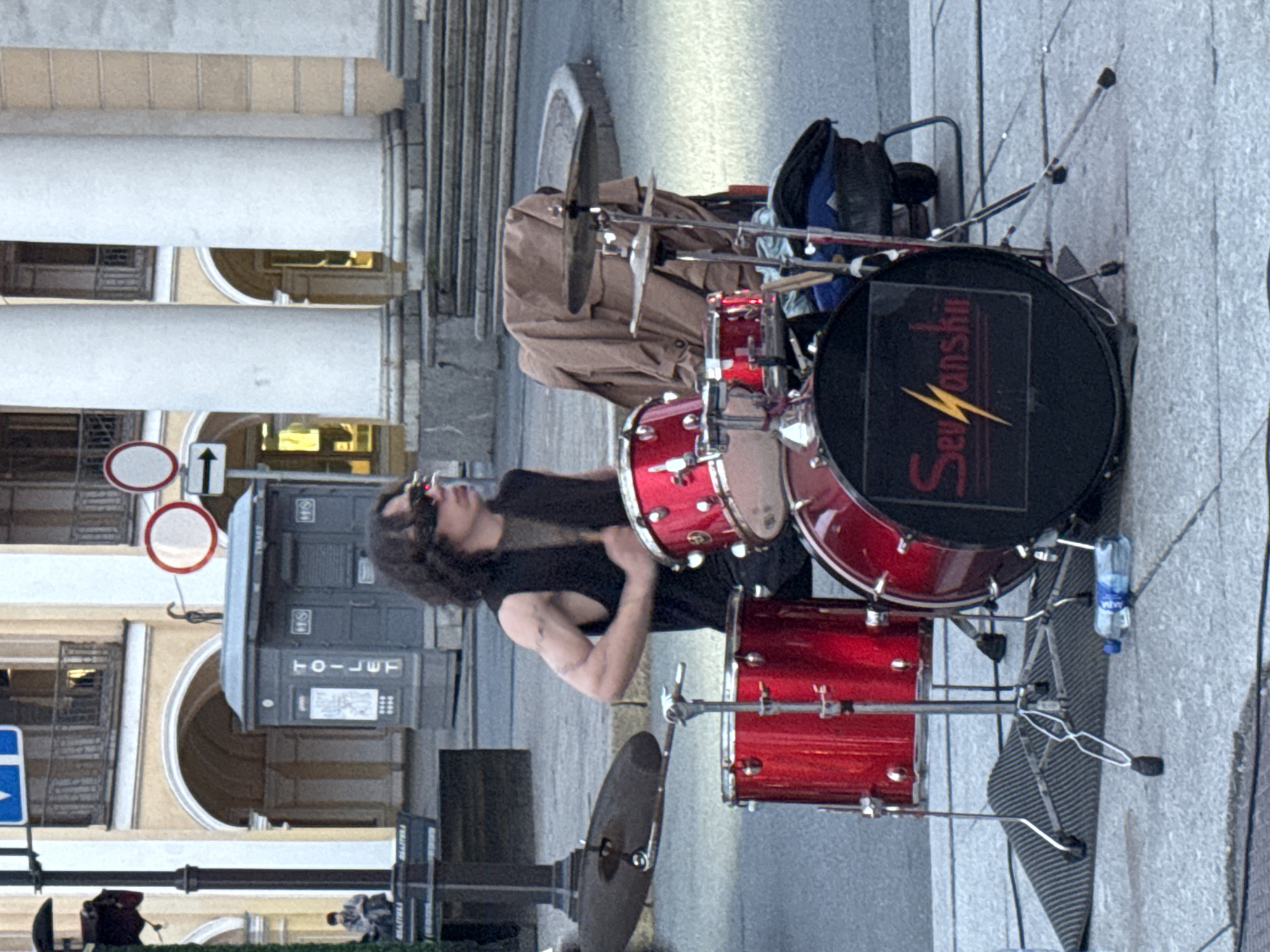 The image depicts a male drummer playing a red drum kit on a paved city street in Saint Petersburg, Russia. The drummer, wearing sunglasses and a black sleeveless shirt, is positioned behind the drum set. His arms are visible, and he appears to be in the process of playing. The drum kit includes a bass drum with a logo that reads "Sevansh" with a lightning bolt symbol, several toms, and cymbals. A brown jacket is draped over a chair behind the drummer. In the background, there is a portable toilet structure with the word "TOILET" visible. Further in the background, classical architecture with large columns and arches is present, suggesting a location like a historical square or street in the city center. Several street signs and a lamppost are also visible. The ground is paved with cobblestones or similar material, and there are some wires and cables on the ground near the drum set. A water bottle lies on the ground next to the drum kit. The overall scene suggests a street performance or rehearsal.