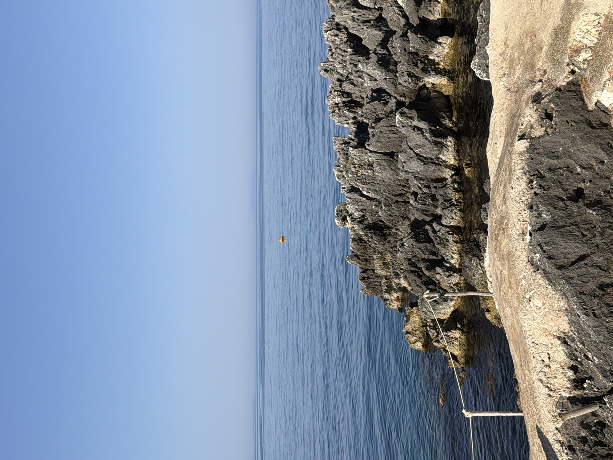 The image depicts a serene coastal scene in Himarë, Albania. The foreground features a rough, light-colored rocky surface, with darker, more jagged rocks forming the middle ground, leading down to the clear, calm blue sea. A thin, yellow rope is strung between two wooden posts in the middle ground, suggesting a path or boundary. In the distance, a solitary yellow buoy floats on the water. The horizon is clearly defined, with a faint outline of land visible on the far left, under a vast, clear, light blue sky. The lighting suggests a bright, sunny day. There are no people or animals visible in the frame.