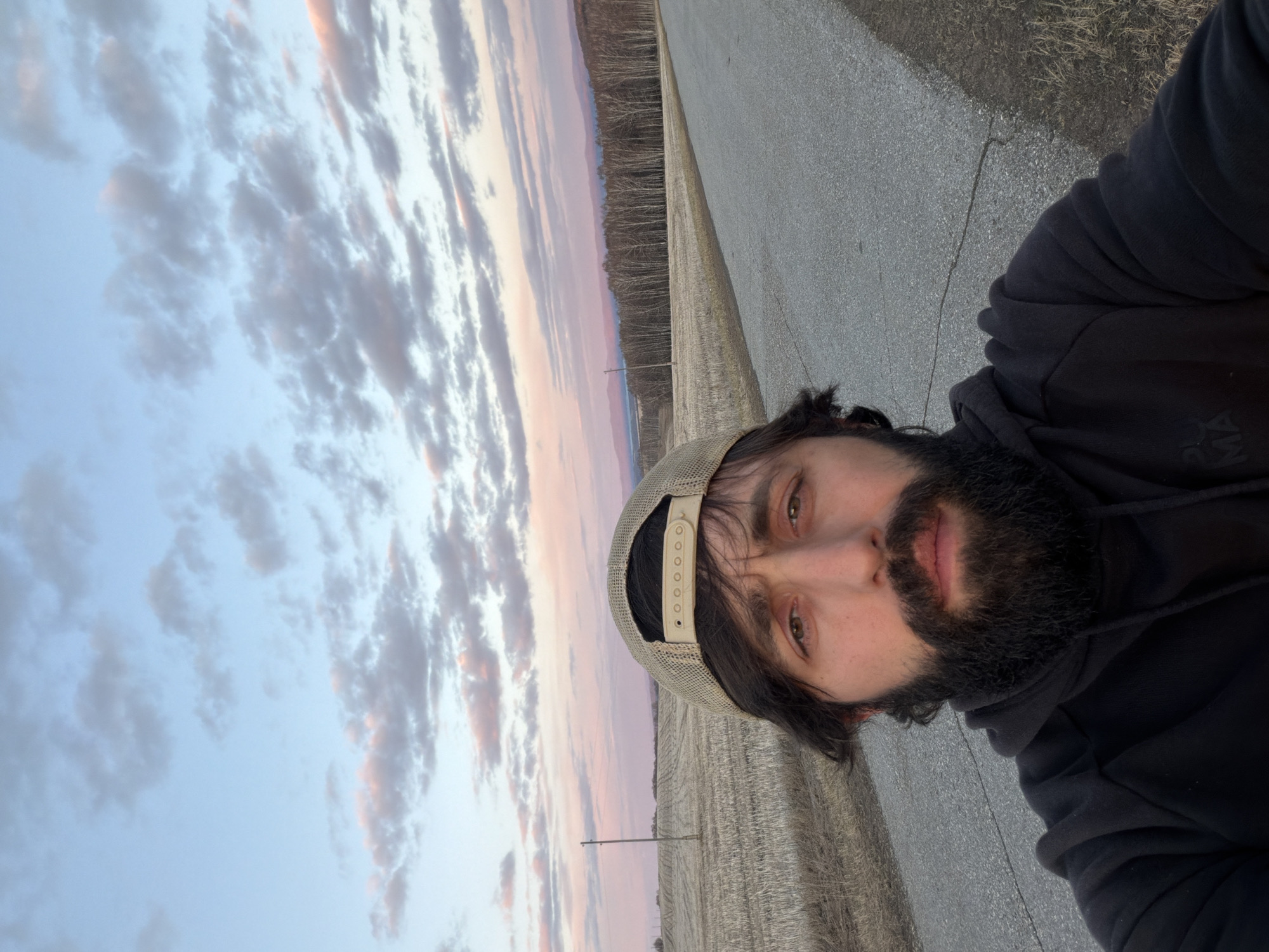 A bearded man wearing a baseball cap and a black hoodie is standing on a gray asphalt surface next to a harvested field. Behind the field, there is a line of bare trees. The sky is filled with clouds colored with hues of pink and blue, indicating either sunrise or sunset.
The location within the city of Chesnoki, Russia, cannot be determined from the visual information provided.
