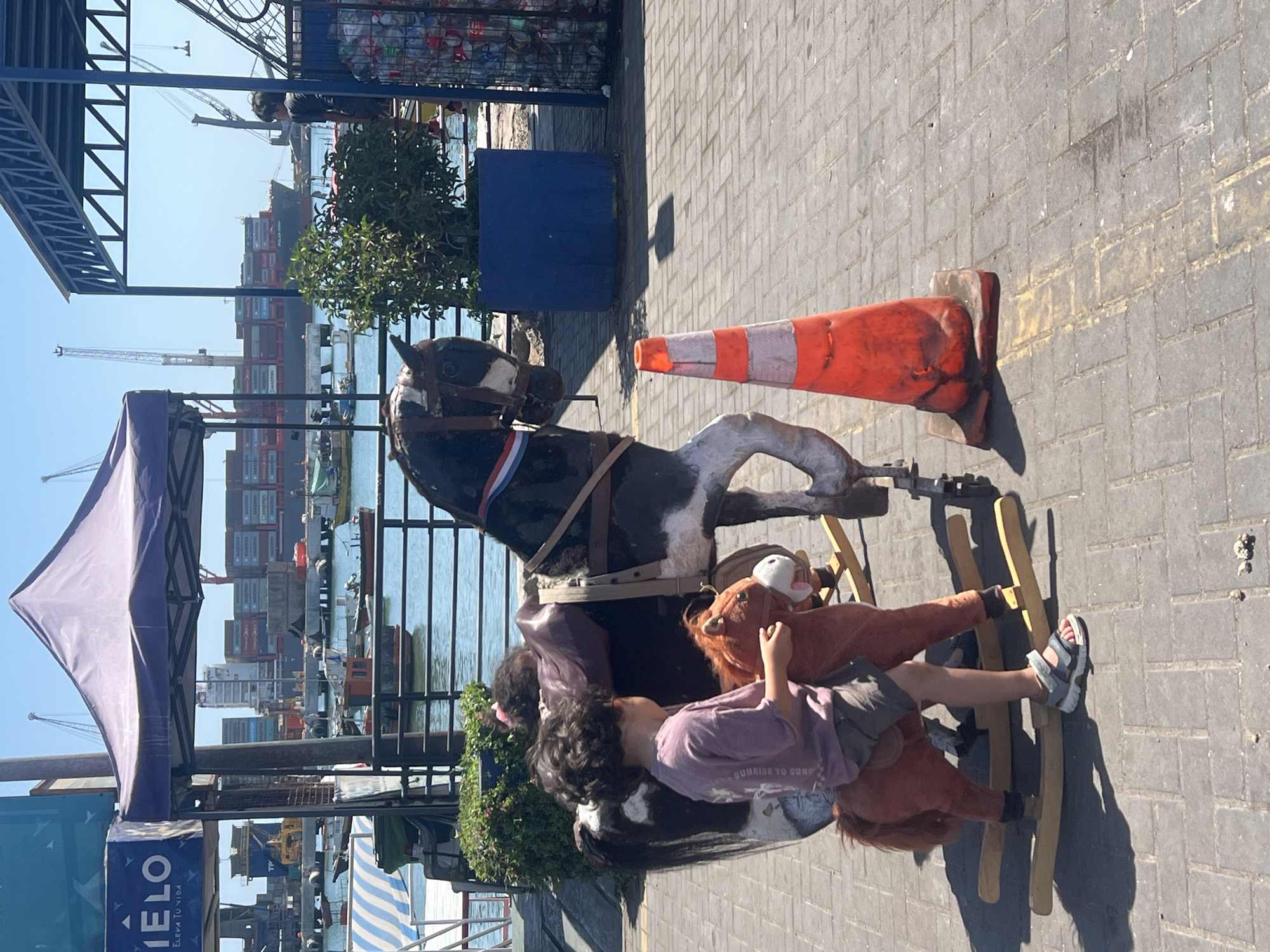 The image captures a street scene in Arica, Chile, featuring a child riding a rocking horse on a brick-paved surface. The child, wearing a purple shirt and gray shorts, is seated on a stuffed horse attached to the rocking mechanism. A larger, black and white rocking horse is positioned adjacent to the child's ride, with an orange traffic cone resting nearby.
In the background, a harbor scene is visible, complete with cargo ships, cranes, and waterfront buildings. A partially visible tent structure with the word "IELO" on it suggests a commercial or recreational area. The scene is bathed in daylight, indicating an outdoor setting.