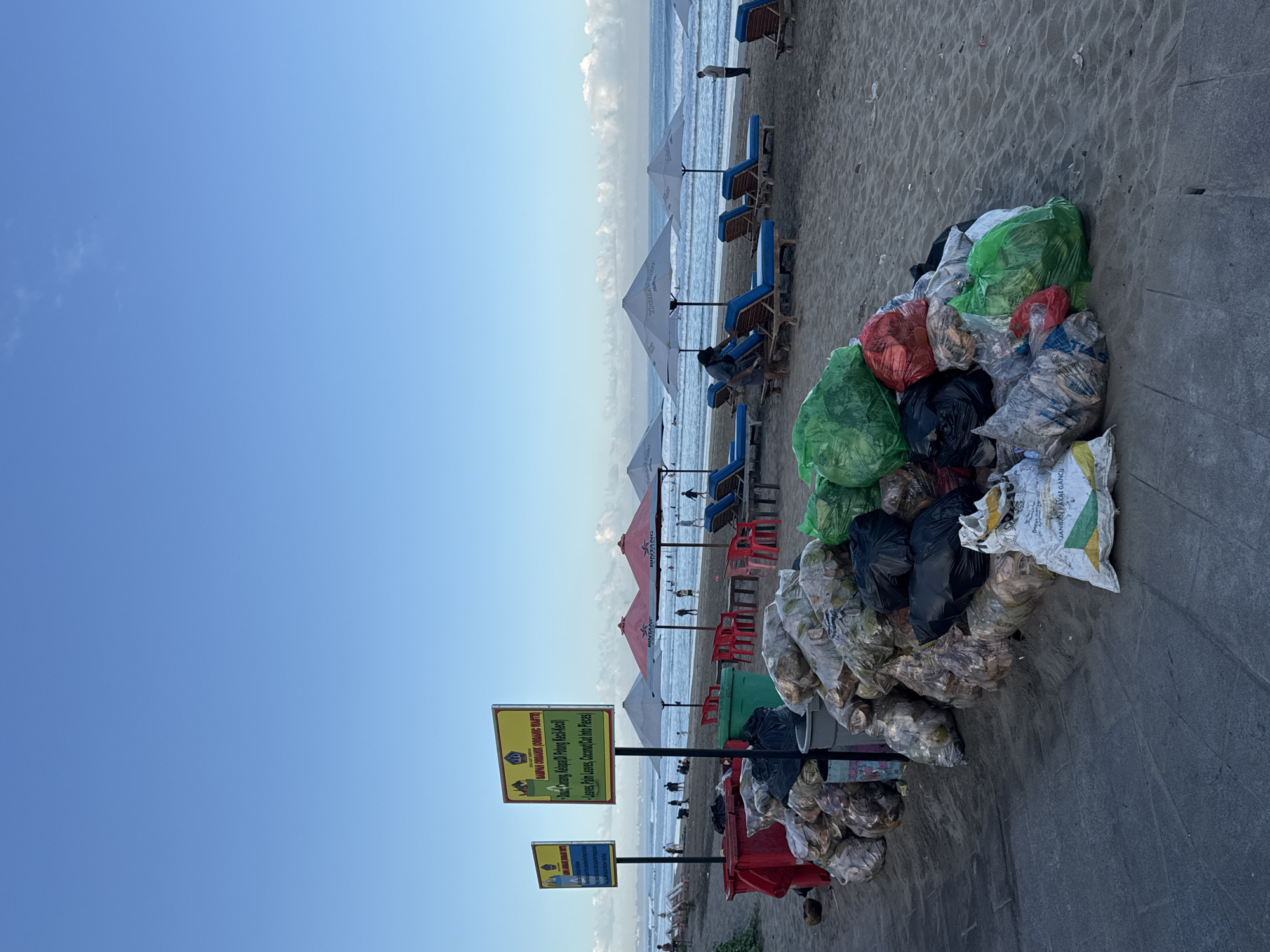 The image depicts a beach scene in Kuta, Indonesia. The beach is lined with lounge chairs and umbrellas. A large pile of garbage bags sits on the edge of the beach near a sign that reads "SAMPAH ORGANIK (ORGANIC WASTE). Leaves, Palm Leaves, Coconut Cut Into Pieces" and another shorter sign beneath, reading "JANGAN PAKAI GANCU" (Do Not Use A Grapple). Several people are visible on the beach and in the water. The sky is blue and the ocean is visible in the background.