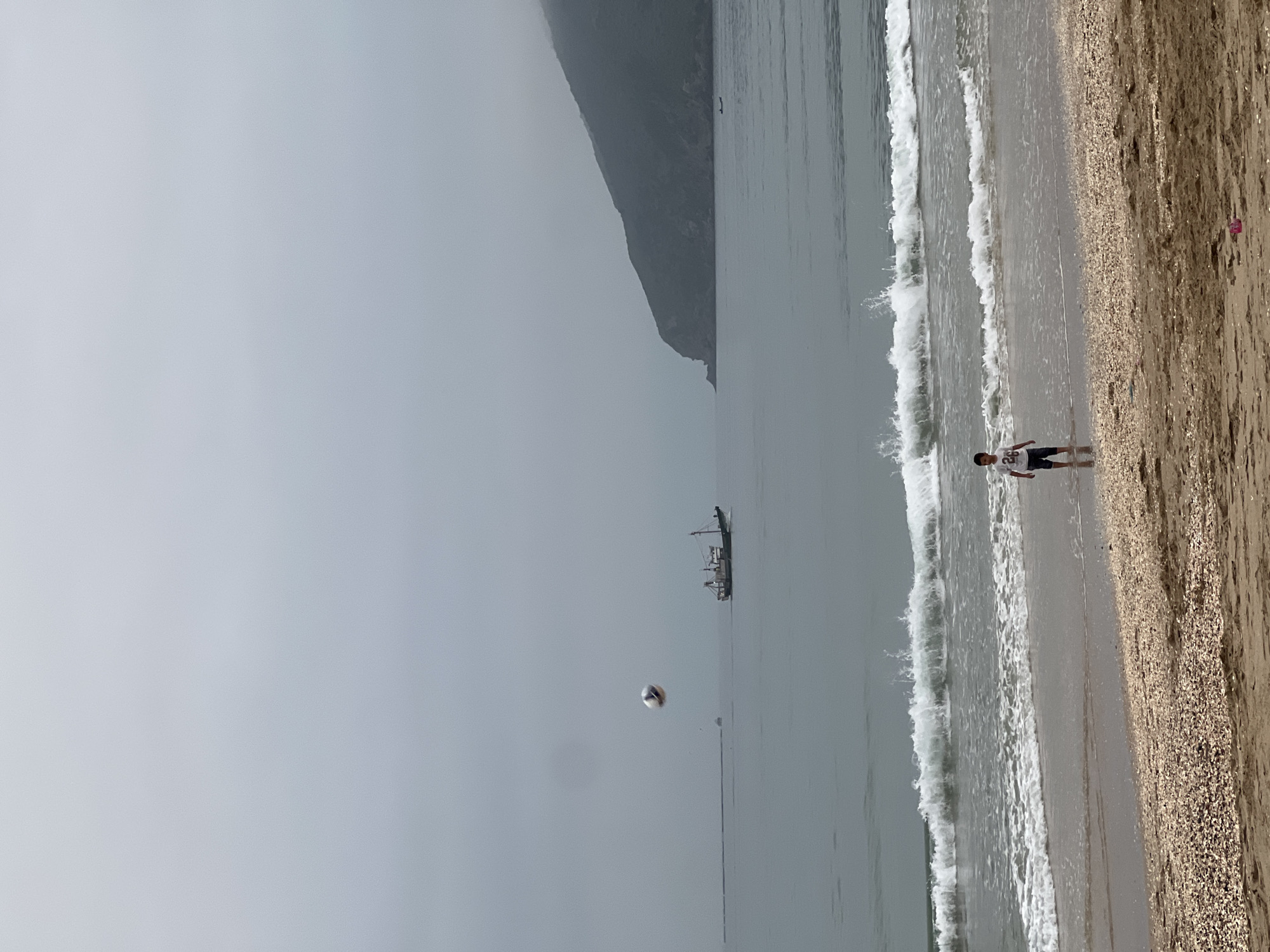 This image captures a hazy daytime beach scene in M'Diq, Morocco. On the left side, a large, dark, and indistinct mountain or cliff rises from the sea, partially obscured by what appears to be overcast or foggy weather. The sky, a soft grey, stretches above the distant landmass and across the upper part of the frame.
The sea occupies the central and right areas of the image, appearing calm with a muted grey tone. In the middle distance, a boat, possibly a fishing trawler, is visible on the water. Closer to the foreground, a white and brown ball, resembling a volleyball, is caught in mid-air above the water, suggesting an ongoing game.
On the far right, the sandy beach meets the sea. Gentle waves with white foam crests break repeatedly on the shore, creating a line between the wet, reflective sand and the drier, tan-colored sand further up the beach. A young boy stands in the shallow water on the right, wearing a white t-shirt with a dark number "25" printed on it and dark shorts. His presence indicates leisurely activity at the beach. The overall atmosphere is tranquil and somewhat muted due to the hazy conditions.
