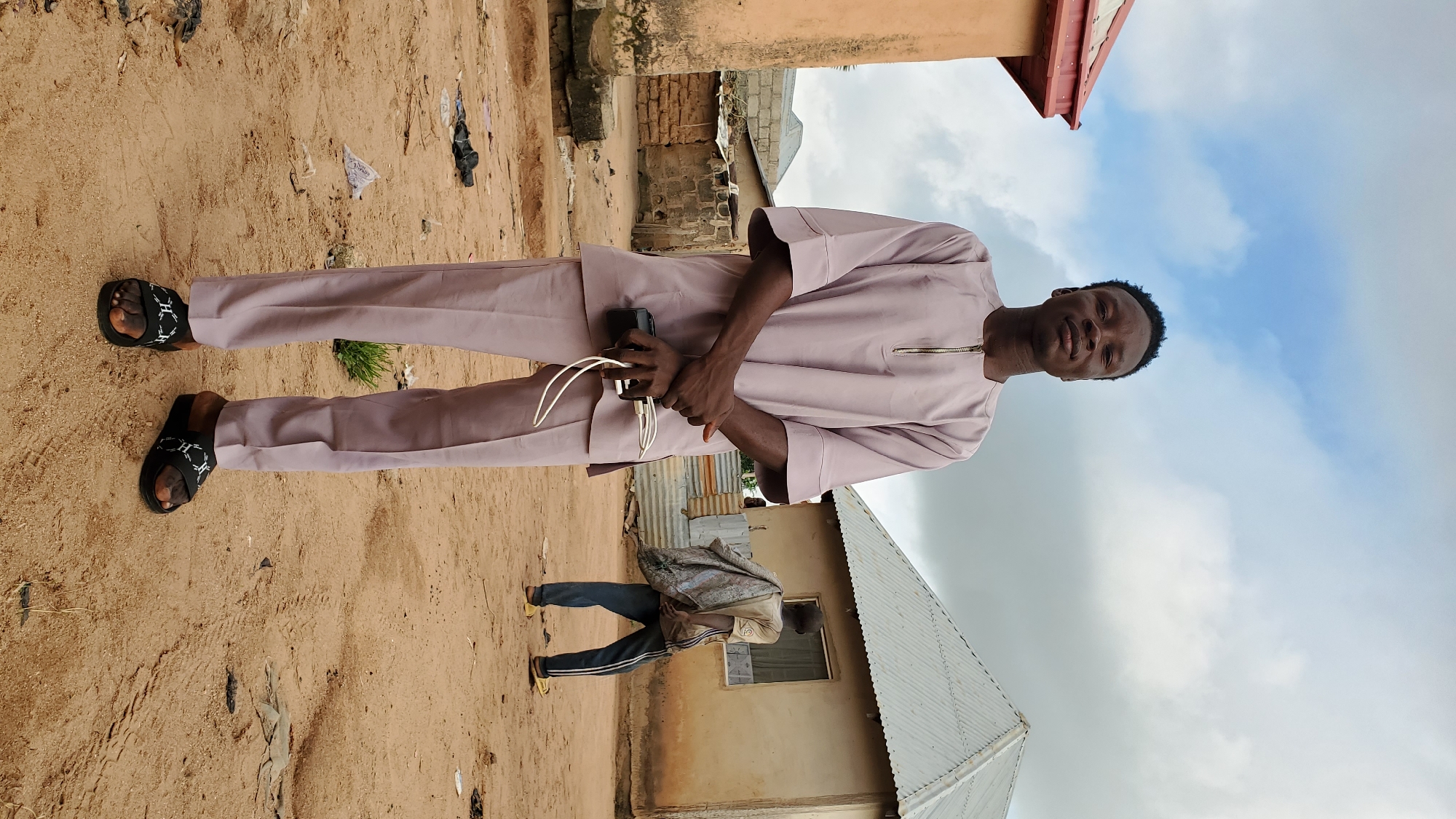 In the city of Unknown, Unknown, a young Black man stands in the foreground, facing the camera. He is wearing a light purple, two-piece traditional outfit with a zipper closure at the neck and loose-fitting pants. His hands are clasped in front of him, and he is holding a black smartphone and white charging cables. He wears black sandals with a white criss-cross pattern. The ground beneath him is sandy and uneven, with scattered debris.
In the background, a second Black man is walking away from the camera, carrying a large, bundled item over his shoulder. He is wearing a light-colored t-shirt and dark pants with white stripes. He is also wearing yellow sandals.
The environment consists of low-rise buildings with corrugated metal roofs and mud-brick walls. The sky is overcast with clouds. The overall scene depicts an outdoor, possibly rural or semi-urban, setting.