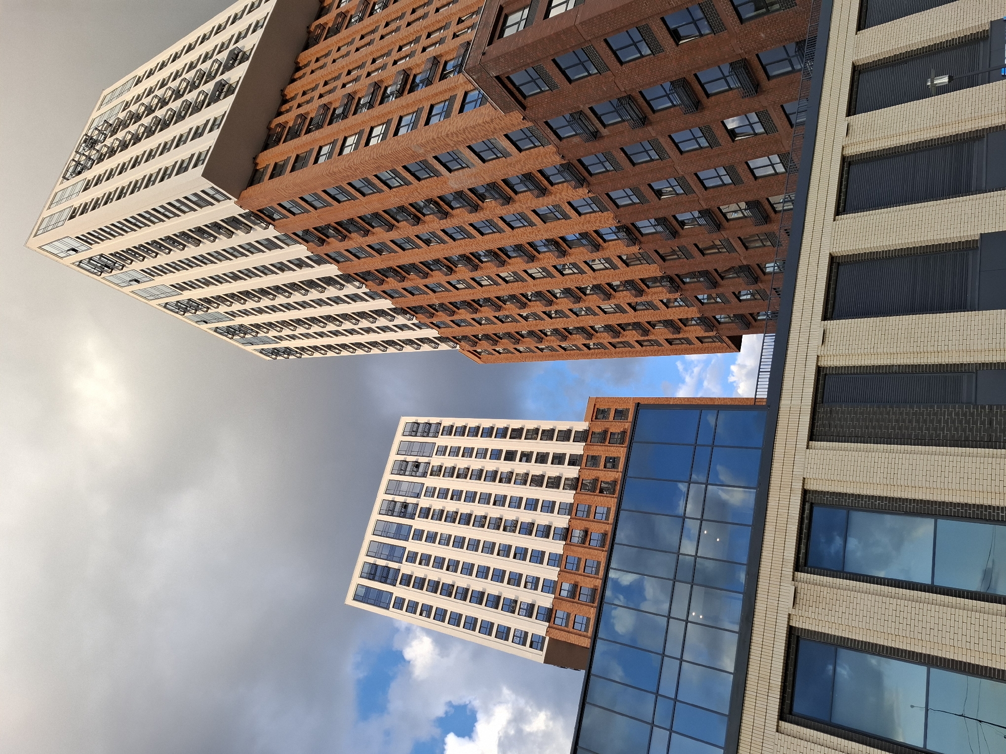 This image captures a low-angle view of two modern high-rise apartment buildings against a partly cloudy sky. The building on the left is primarily clad in light-colored panels with numerous windows, sitting atop a section of red brick and a lower structure with large, reflective glass windows. The building on the right is taller and features a design with a red brick lower section and a white upper section, also with many windows and projecting balconies. The style of architecture suggests a contemporary residential complex. The image is taken from a ground-level perspective looking upwards, emphasizing the scale and modern design of the buildings. The lighting suggests it is daytime, possibly late afternoon given the direction of the light and cloud cover. The scene is devoid of any people or vehicles. This particular complex, with its distinctive architecture, is located in the city of Tyumen, Russia, and is known as the "Toshno" residential complex, situated on Gorkogo Street.