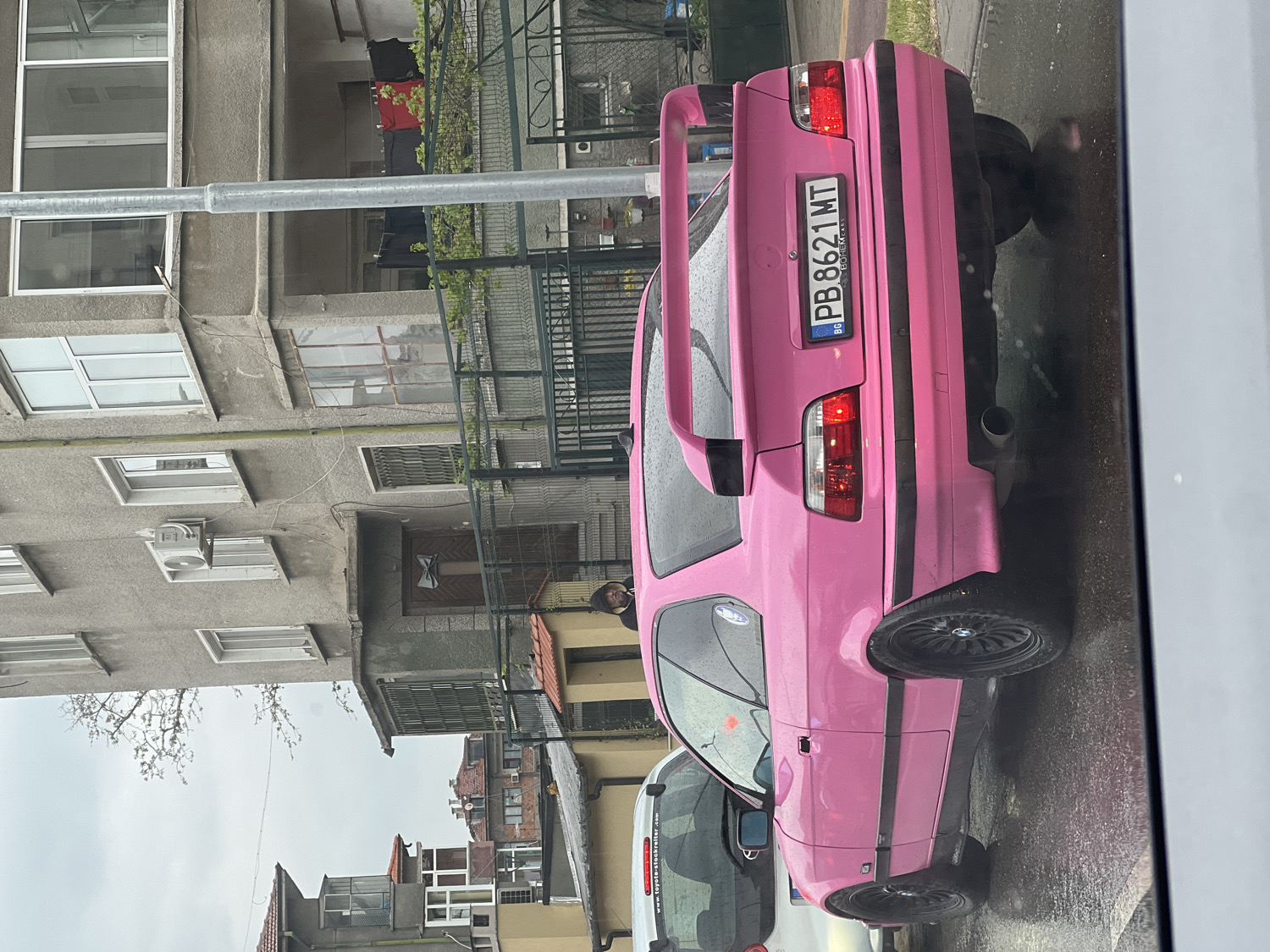 A vibrant, custom-painted pink BMW, likely an E36 model coupe, is the central focus of this image, parked on a wet street in what is identified as Plovdiv, Bulgaria. The car stands out with its striking color, a large, color-matched rear spoiler, and black multi-spoke alloy wheels. A prominent Bulgarian license plate, "PB 8621 MT," is affixed to the rear, with "PB" indicating the Plovdiv region. A single exhaust pipe is visible below the rear bumper.
The scene is set against a backdrop of multi-story residential buildings, characterized by their grey facades, various window styles, and several balconies. One building section in the background features clothes drying on a clothesline, and an external air conditioning unit is mounted on another part of the building. The ground appears wet, suggesting recent rain under an overcast sky, indicating it is daytime.
A person in a dark jacket is partially visible near the rear of the pink car. To the left (or front in an upright view) of the pink BMW, a portion of a white commercial vehicle is seen, bearing the website address "www.toyota-forklift.bg," providing an additional location cue. The overall atmosphere depicts an ordinary urban street scene, made distinctive by the unusually colored vehicle.
