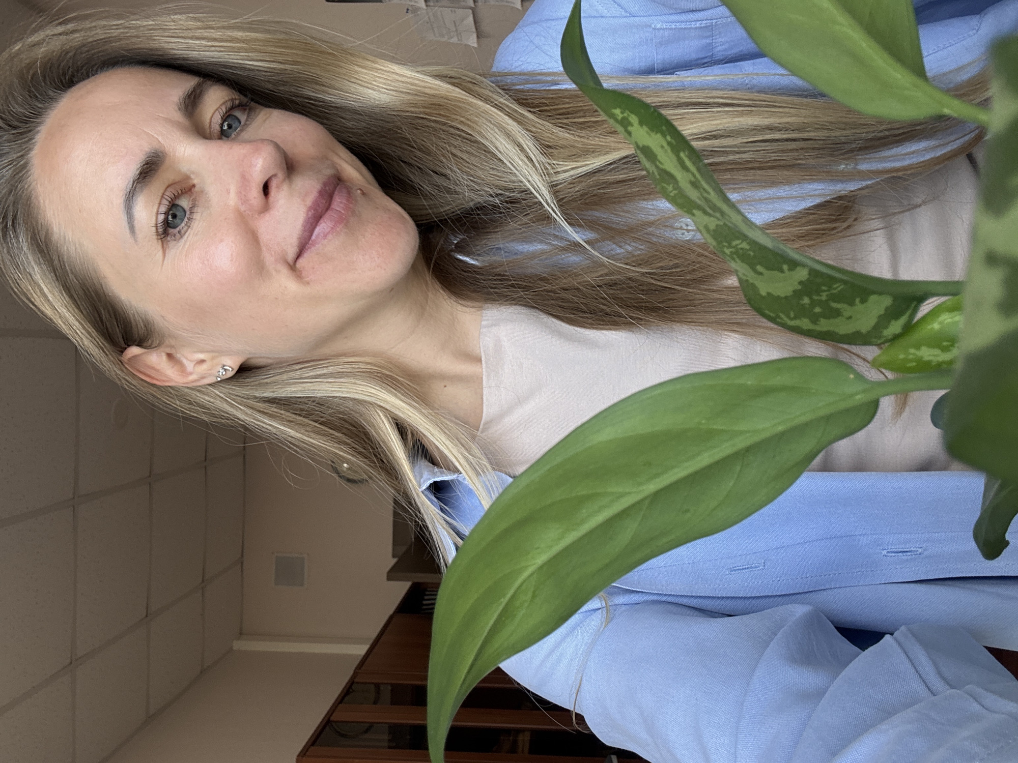 A woman with blonde hair and blue eyes is taking a selfie indoors. She is wearing a light blue button-down shirt over a beige top and a small silver earring in her left ear. In the foreground, the large green leaves of a houseplant partially obscure the woman and the background. The environment appears to be an office or a room with a drop ceiling, bookshelves, and what looks like a radiator. There is no visible indication of the location being specifically in the city of Chaikovsky, Russia.