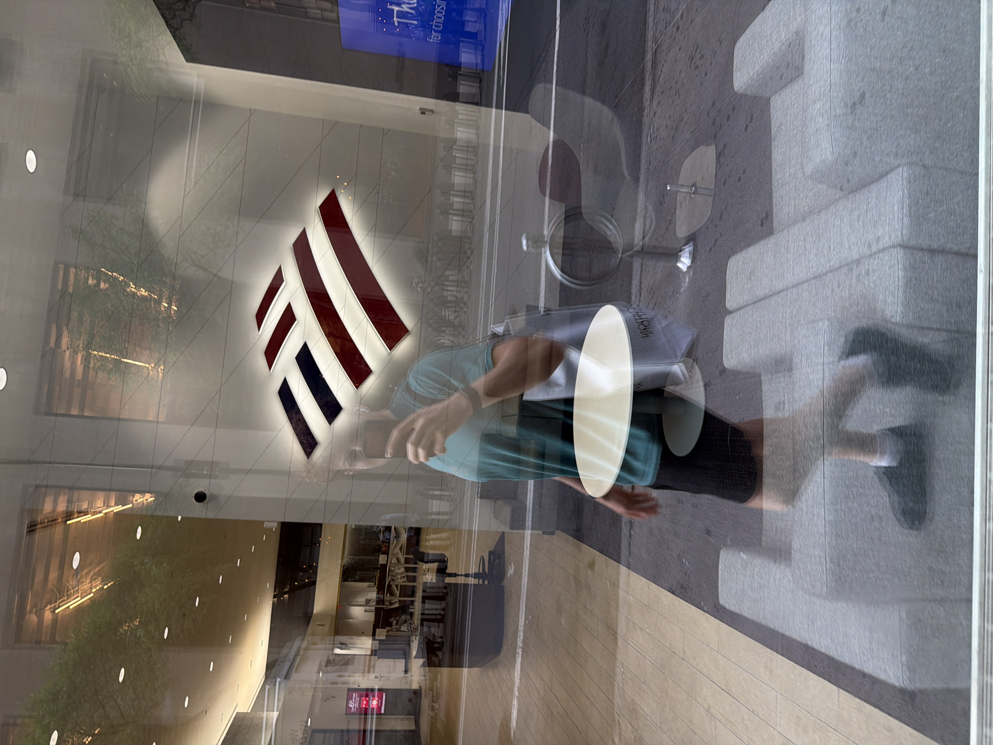 The image is a reflection in a Bank of America building's window. A person carrying a white, cylindrical object is visible. The reflection shows an outside street scene, including a curb and pedestrian walkway. A blue sign hangs in the upper portion of the window. The branch’s illuminated logo is a prominent feature in the reflection. Inside the bank, a seating area with furniture can be seen, along with a security camera. The location is likely in New York City based on the Bank of America logo.