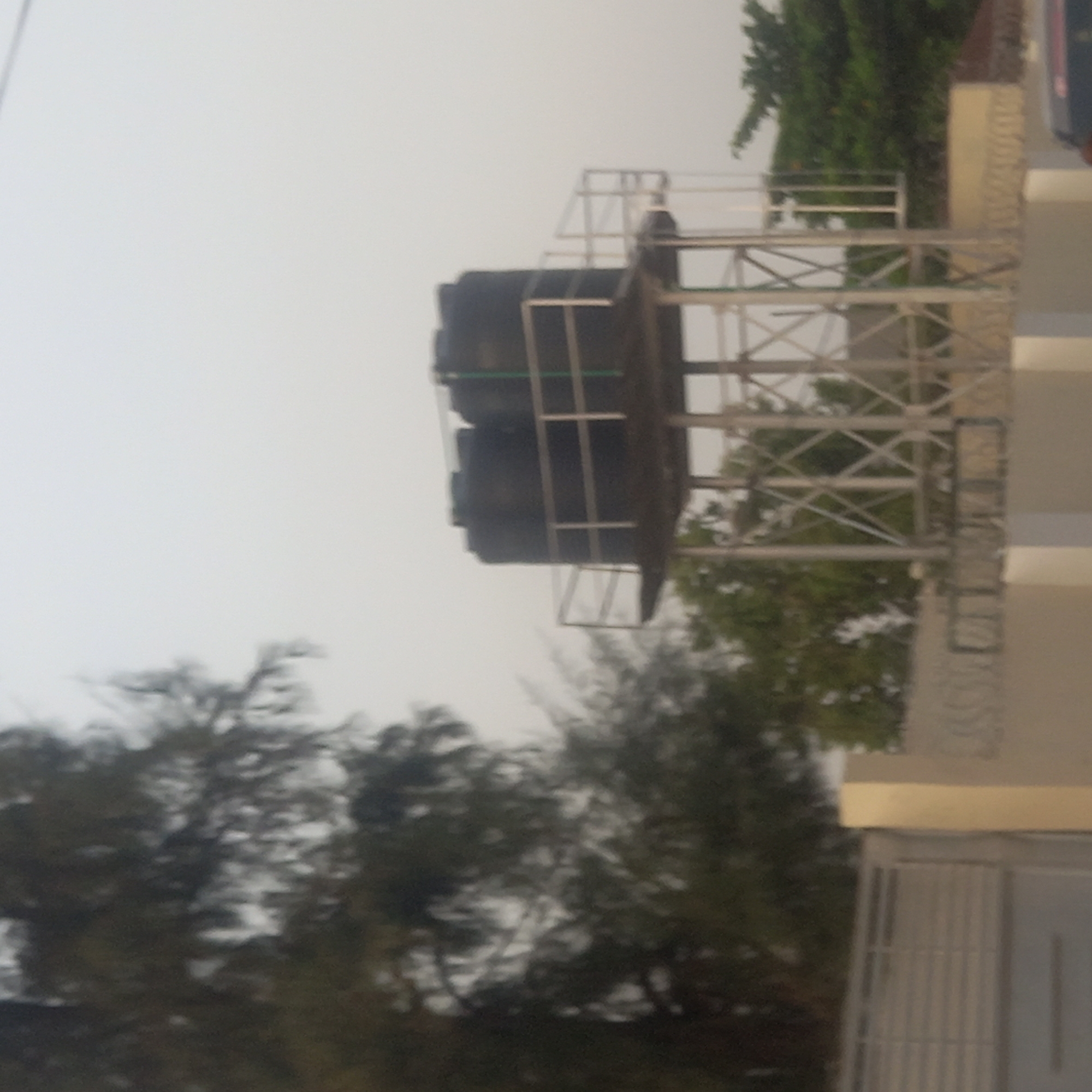 The image shows two black water tanks on a metal platform, likely located in Dadi Riba, Nigeria. The platform is constructed with a silver, lattice-like frame and surrounded by a railing. The tanks are positioned on top of the platform. To the left, there are blurred tree branches and foliage. To the right, a building with a tan facade is partially visible, including a metal gate or door. The sky is overcast, casting a diffused light. The angle suggests the photo was taken from a low position.