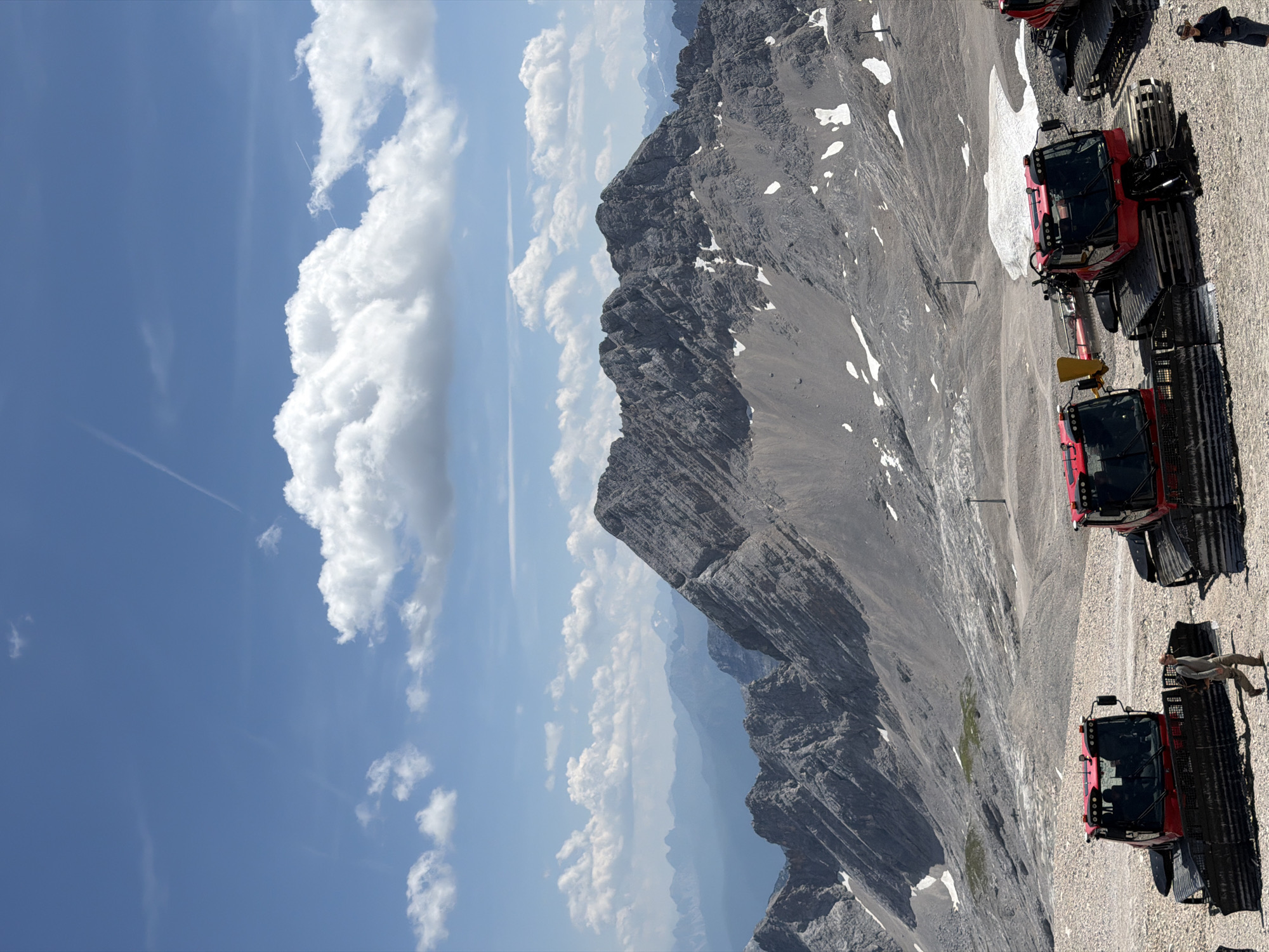 The image shows a bright, sunny day in the Bavarian Alps, likely near the Zugspitze, Germany's highest peak, which is accessible from Garmisch-Partenkirchen. In the foreground, three red snow groomer vehicles are parked on a rocky, gravelly terrain. One vehicle on the left is partially visible, while the other two are more prominent. A man in a light-colored shirt and pants walks between the two main snow groomers. In the background, imposing, rugged mountains rise under a vivid blue sky dotted with white, fluffy clouds and contrails. Patches of snow are visible on the mountainsides, indicating high altitude. The scene conveys a sense of a remote, mountainous environment, possibly a ski resort area during the summer season or early autumn.