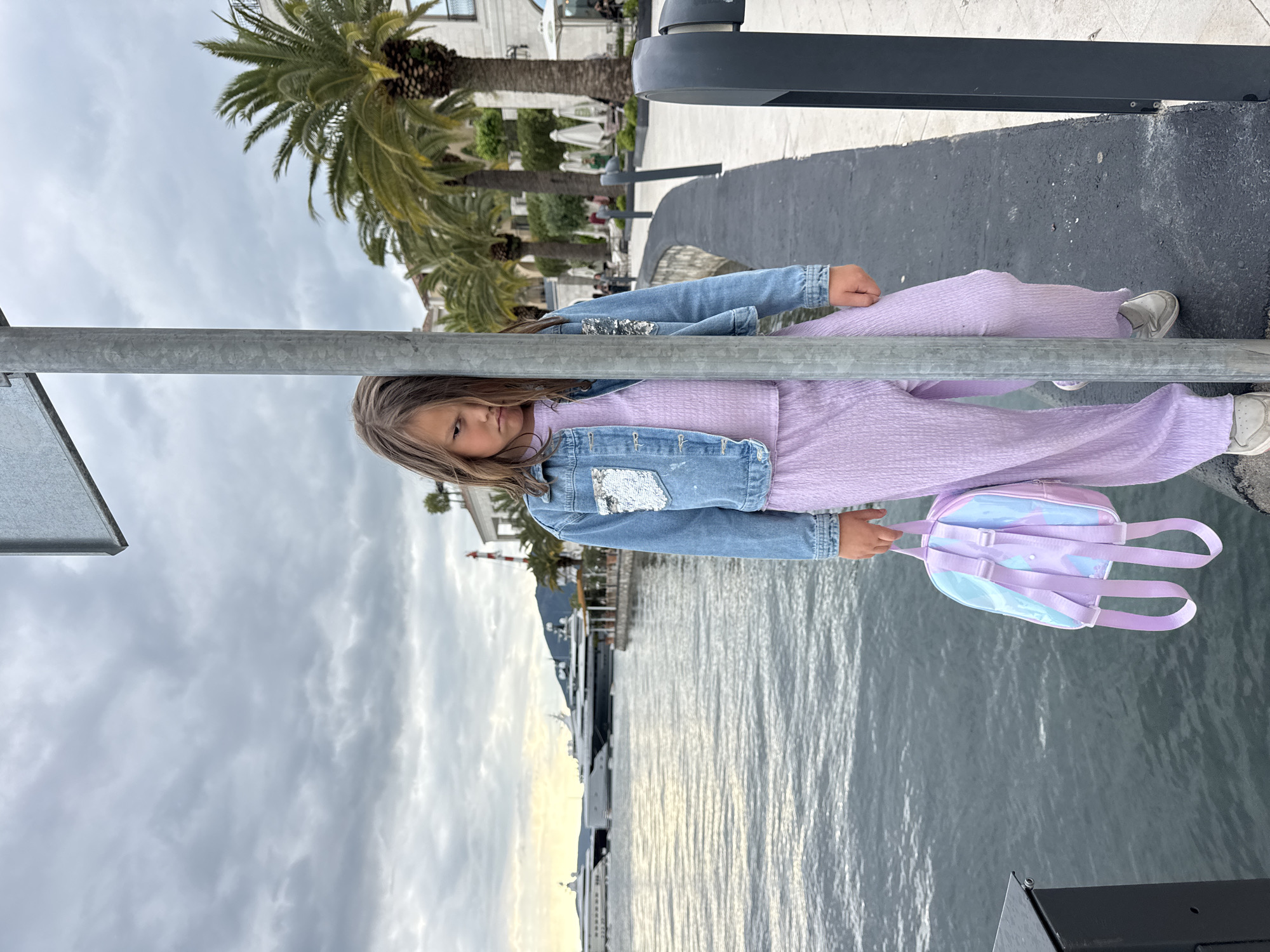 A young girl stands on a gray paved area, possibly a walkway or pier, in Tivat, Montenegro. She wears a lavender jumpsuit with a denim jacket featuring sequined patches and gray sneakers, and she is holding a small purple backpack. A gray metal pole intersects the foreground of the image. Behind the girl, the water of the bay stretches into the distance with a backdrop of cloudy sky. Buildings, palm trees, and distant mountains are visible on the opposite shore, along with boats docked in the water.