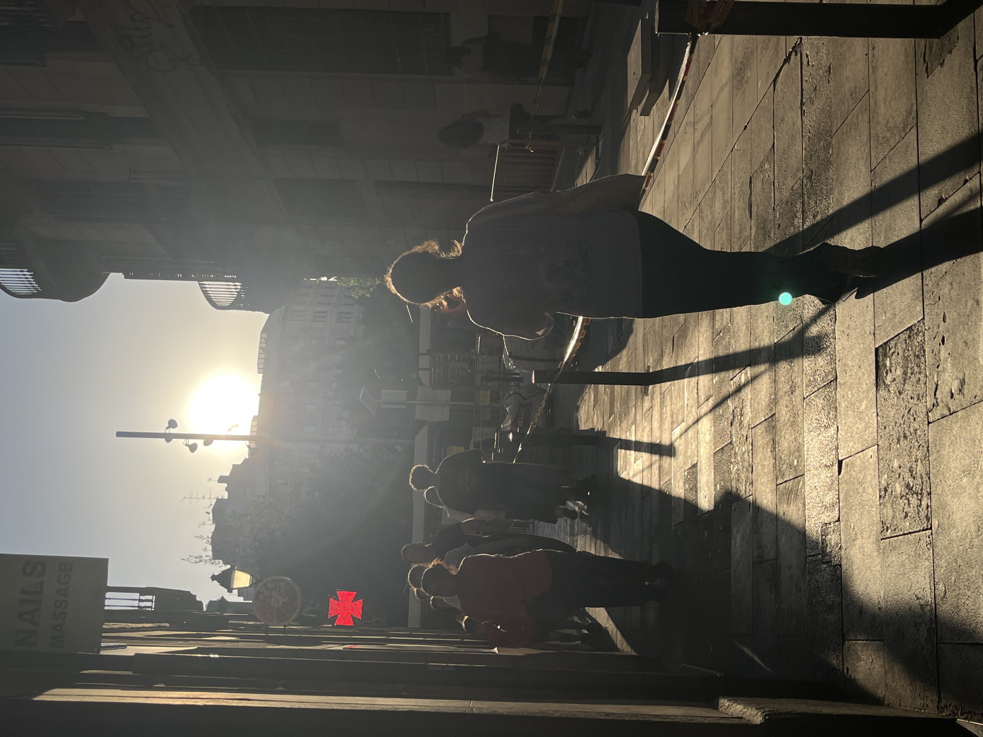 The image captures a street scene in an unidentified city, possibly during the early morning or late afternoon given the position of the sun in the sky. The location appears to be a pedestrian area paved with stone blocks. Several people are walking along the sidewalk, silhouetted against the bright sunlight. The silhouettes indicate a range of body types and clothing styles. A sign is visible on the left edge of the frame, partially obscured by shadow, displaying the words "NAILS MASSAGE." A red cross emblem is also visible in the lower left quadrant. The buildings in the background are generic urban structures. The street is bathed in strong sunlight, creating deep shadows and high contrast.
