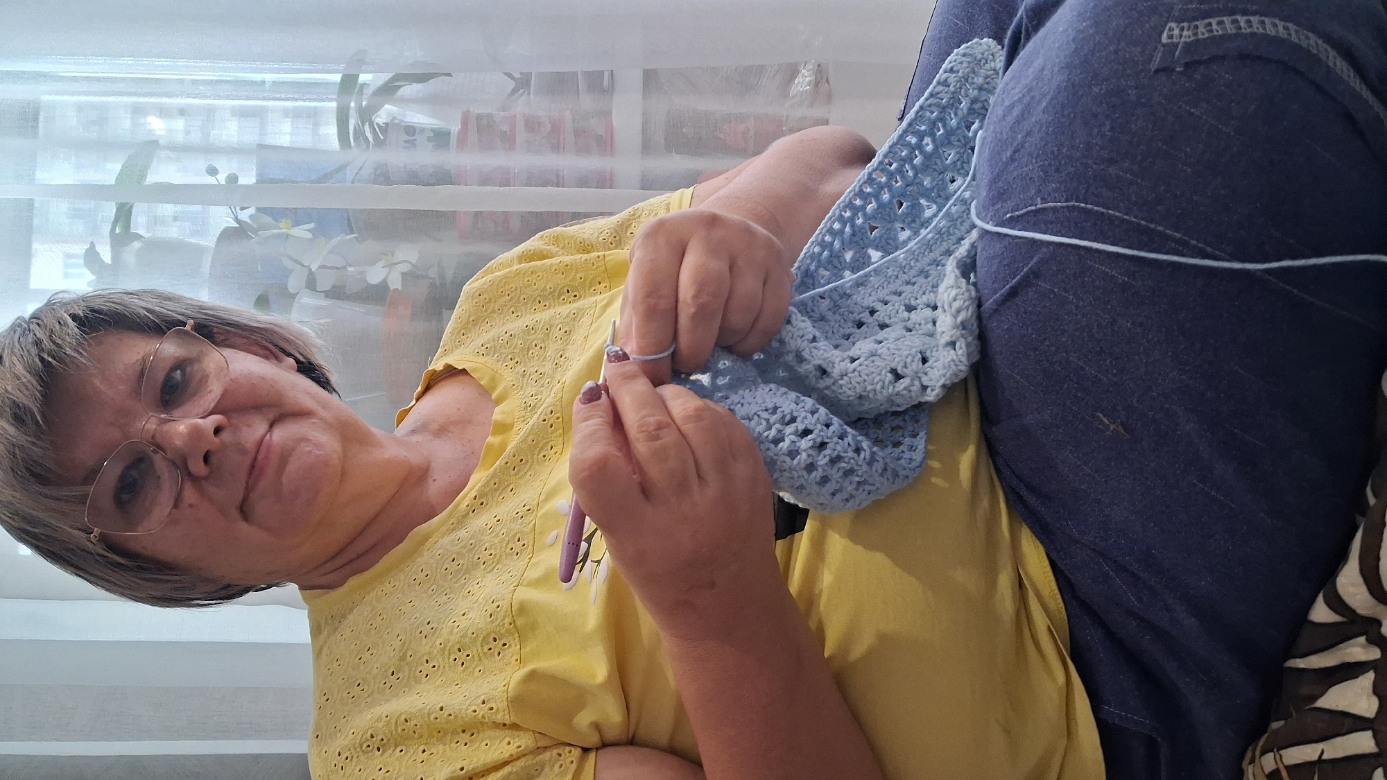 A woman with short, graying hair and glasses is sitting and crocheting with light blue yarn. She is wearing a yellow t-shirt with a patterned neckline and dark blue pants. The background shows a window with white curtains, and some plants are visible. The scene appears to be indoors, likely in a residential setting in Koltsovo, Russia.