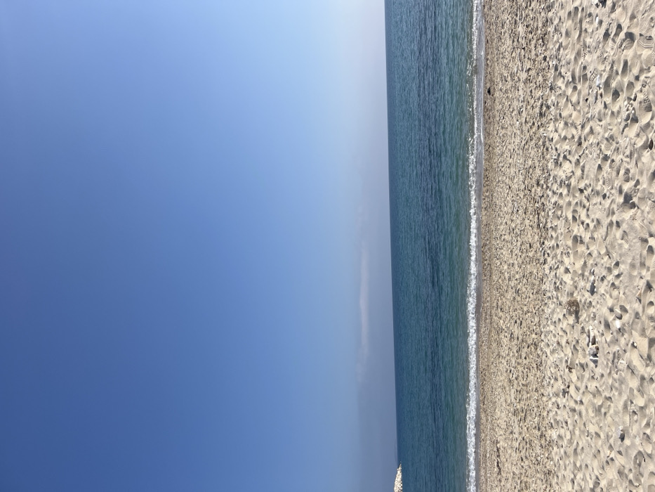 The image shows a wide, sandy beach in Limenaria, Greece, at coordinates 40.626, 24.571. The beach is empty, with no people visible. There are a few footprints in the sand, and a small rocky outcropping visible in the distance. The water is a clear blue and the sky is cloudless. There is a small wave crashing onto the shore in the foreground. The image is taken from a low angle, looking out towards the sea.