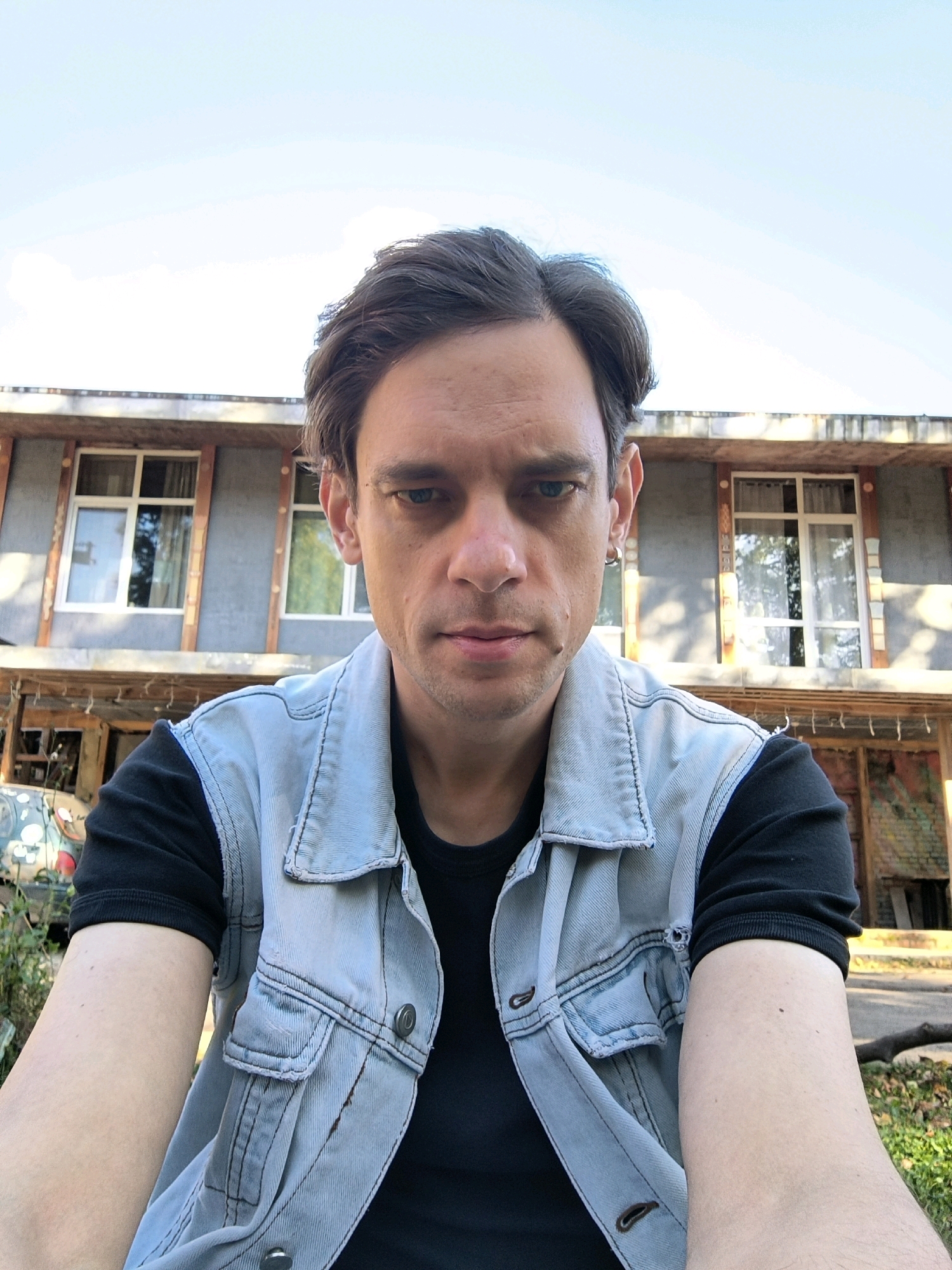 A man, appearing to be in his late 20s to early 40s, is captured in a selfie taken outdoors. He has brown hair, styled casually, and is looking directly at the camera with a neutral expression. His skin tone is fair, and he has faint stubble on his chin and jawline. He is wearing a black t-shirt underneath a light blue denim vest. The vest has prominent stitching and two buttoned pockets on the chest.
Behind him, a two-story building is visible. The building's facade appears to be grey with wooden accents around two visible windows on the upper level. The windows have white frames and are partially obscured by curtains. The lower part of the building shows some exposed wooden beams and what looks like a sheltered area with some graffiti art. The overall setting suggests a somewhat rustic or older urban environment. The lighting indicates it is daytime, with natural light illuminating the scene, casting subtle shadows. The background greenery and the natural light suggest the photo was taken in a temperate climate. The presence of a building and greenery places the scene likely within a populated area, possibly residential or in a less commercial district of Il'inskiy Pogost, Russia.