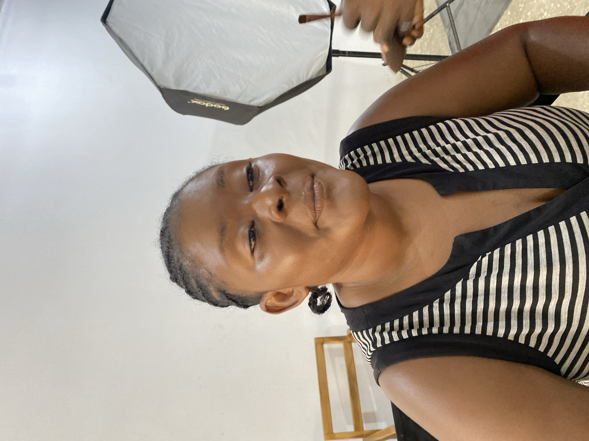 A medium close-up shot of a woman with dark skin wearing a black and white striped top, turned slightly to the side. Her hair is braided back from her face. Above her is a large, octagonal softbox light. The background is a white wall. At the bottom of the frame, there's a partial view of a wooden frame or structure. An arm holding a makeup brush or tool is visible at the top right. The photo appears to be taken indoors. There is no information in the photo to support the location in Gura Top, Nigeria.