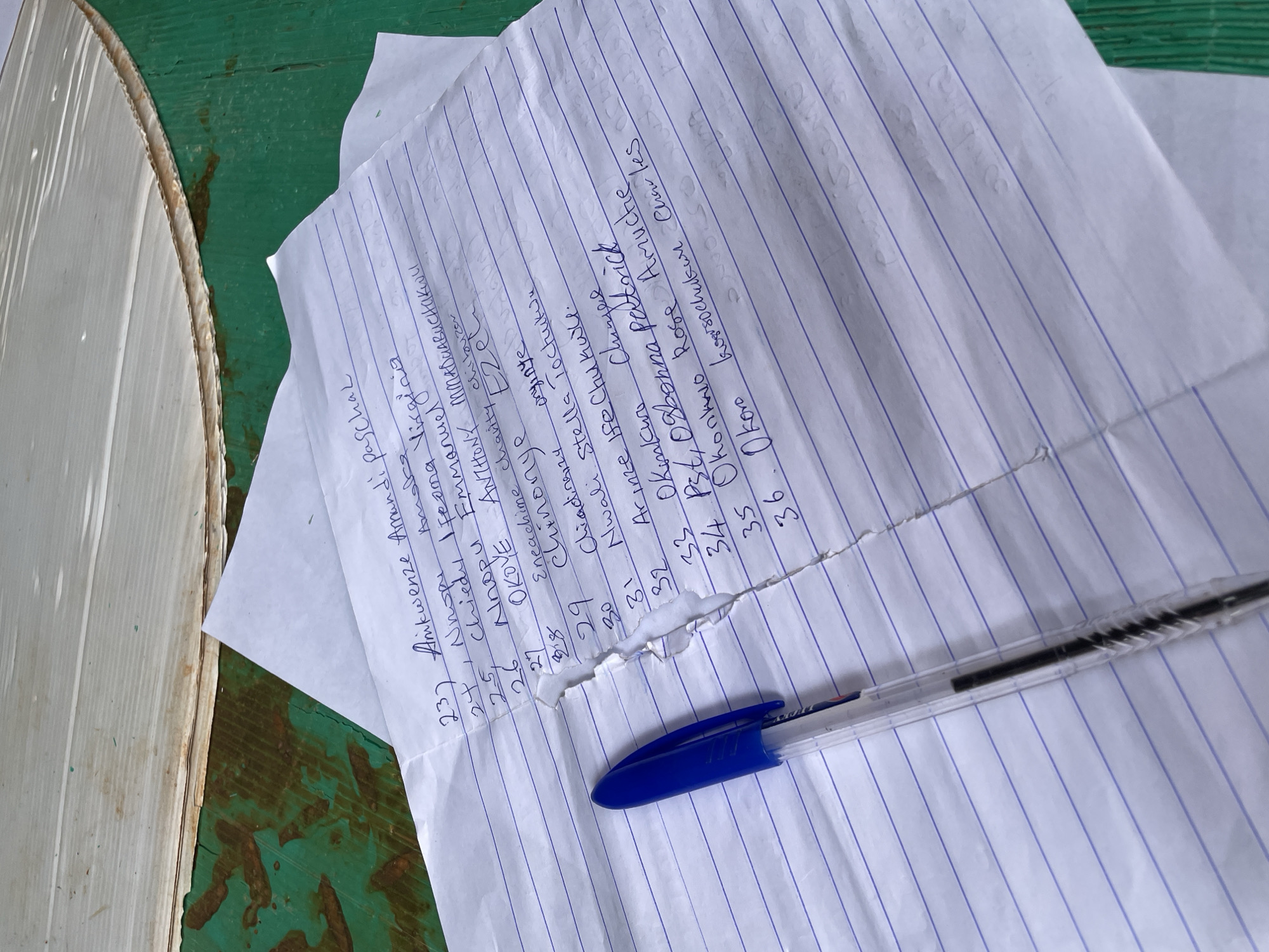 This media file features a close-up view of a handwritten list on lined paper, likely a roster or attendance sheet. The writing, done in blue ink, appears to be in a Nigerian dialect, specifically referencing "Ogidi, Nigeria" in the location context. The paper is slightly crumpled and torn, suggesting it has been handled. A blue ballpoint pen rests on the paper, poised as if in the middle of an entry.
The list contains numbered entries from 23 to 36, with names and some additional notes. The names appear to be a mix of personal names and possibly titles. Some entries, like "27 OKOVE ANTHONY MMADUABUCHakoll" and "34 Pst, Ogbonna patrick," are quite long. The handwriting is somewhat variable in legibility, with some letters forming loops and others appearing more angular. The paper itself is creased and has a visible tear between entries 28 and 31, indicating its use and potential age.
The background shows a textured, green surface, possibly a wooden table with peeling paint, and a portion of a light-colored, textured object, perhaps a woven mat or a drum. The lighting suggests it could be daytime, with natural light illuminating the scene. The overall impression is one of a practical, everyday document in use. The presence of names and numbers suggests an organized listing, possibly for an event, membership, or record-keeping in Ogidi, Nigeria.