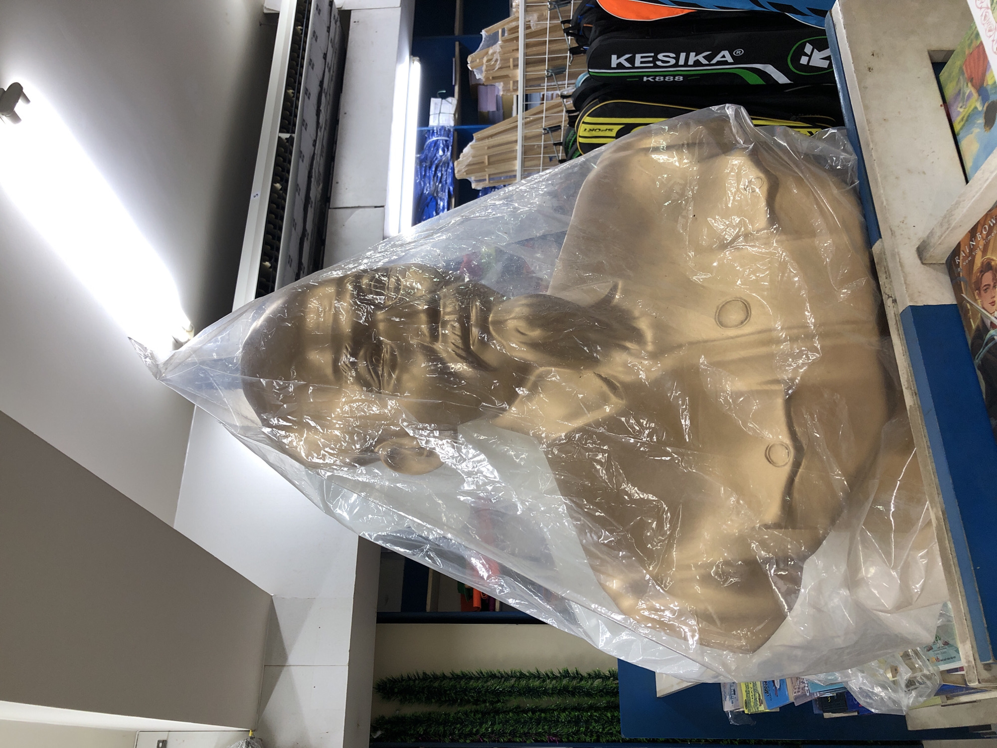 The image shows a golden bust of a man, covered in plastic, displayed in a store in Da Nang, Vietnam. The bust is life-sized and appears to be made of metal or a similar material. It is positioned in the foreground, with its head and shoulders visible. The plastic covering is transparent and crinkled, obscuring some of the details of the bust.
The background of the image shows shelves filled with various items, including what appear to be books, sports equipment bags, and other merchandise. A fluorescent light fixture is visible on the ceiling, illuminating the store. There are no people visible in the image. The overall impression is that of a retail environment, possibly a souvenir shop or a general store, in Da Nang, Vietnam.