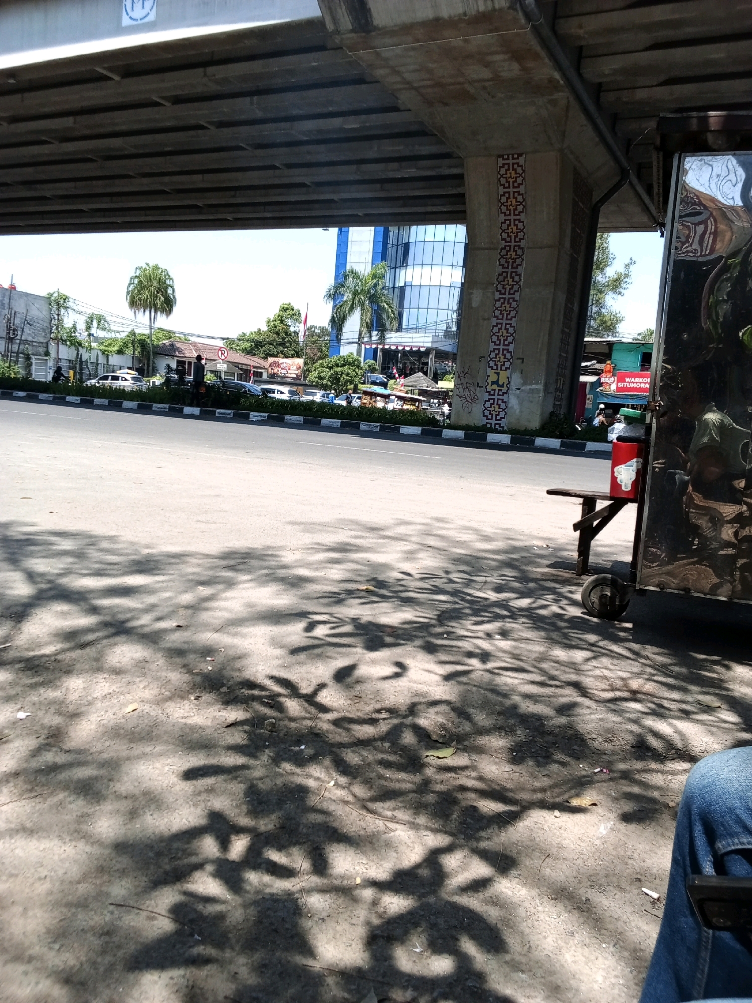 A low-angle, outdoor shot depicts a street scene in Bandung City, Indonesia, under an elevated road structure. The foreground is dominated by a cracked asphalt ground, with dappled shadows of leaves cast across it, suggesting a sunny day with trees nearby. To the right, a portion of a person's legs, clad in blue jeans, is visible, seated on what appears to be a bench. Next to the bench is a wheeled cart with a shiny, reflective surface, possibly a food or beverage vendor's stall. A red plastic container with a green lid sits on the cart. The midground shows a paved road with traffic, including several cars and motorcycles. The road is bordered by a median strip with black and white markings. In the background, buildings are visible, including a modern glass-fronted building and smaller, traditional houses. A red and white Indonesian flag flies from a pole. The scene is framed by the concrete pillars and underside of the elevated roadway, which features a decorative tile pattern on one of the pillars.