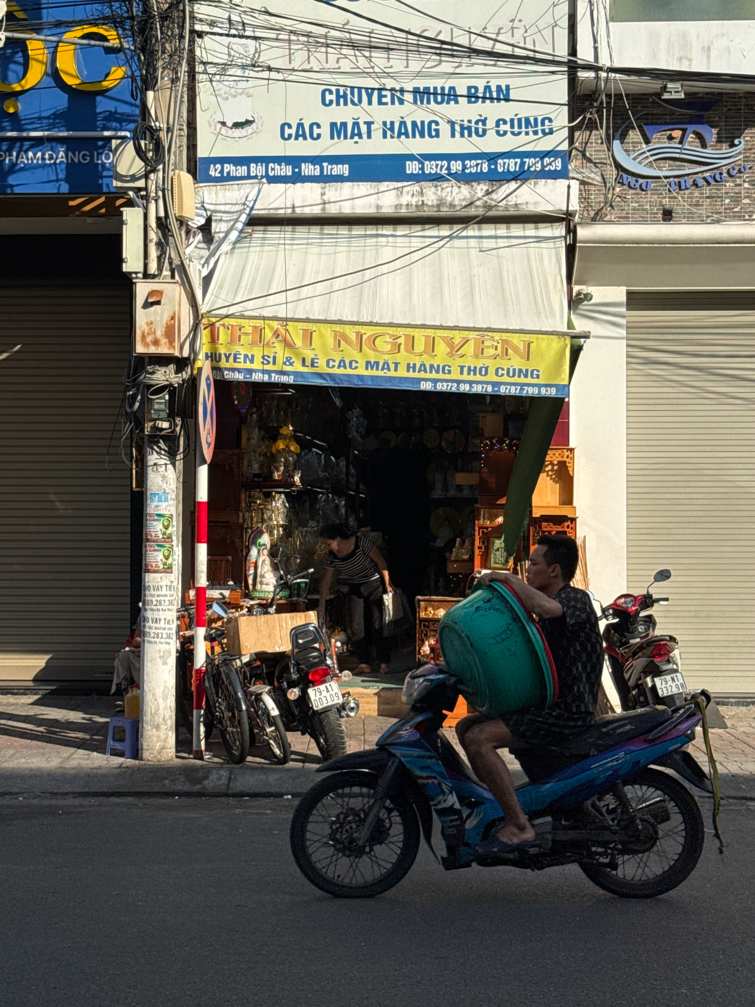 The image shows a street scene in Nha Trang, Vietnam, with a focus on a shop and a man on a motorcycle. A shop with a yellow awning, labeled "THÁI NGUYÊN HUYỀN SĨ & LẺ CÁC MẶT HÀNG THỜ CÚNG" and also "CHUYÊN MUA BÁN CÁC MẶT HÀNG THỜ CÚNG," is visible. The shop's address is indicated as "42 Phan Bội Châu - Nha Trang." Inside the shop, shelves are filled with religious or ceremonial items.
A man is riding a blue and purple motorcycle, carrying a large green tub balanced on his lap. He is wearing a dark patterned shirt and dark shorts. A red and white striped pole stands on the sidewalk near the shop, with various posters and notices attached to it. In the background, other motorcycles are parked, and a person is partially visible inside the shop. The street is paved, and the buildings have corrugated metal roll-up doors. The lighting suggests it is daytime, with shadows cast on the ground.