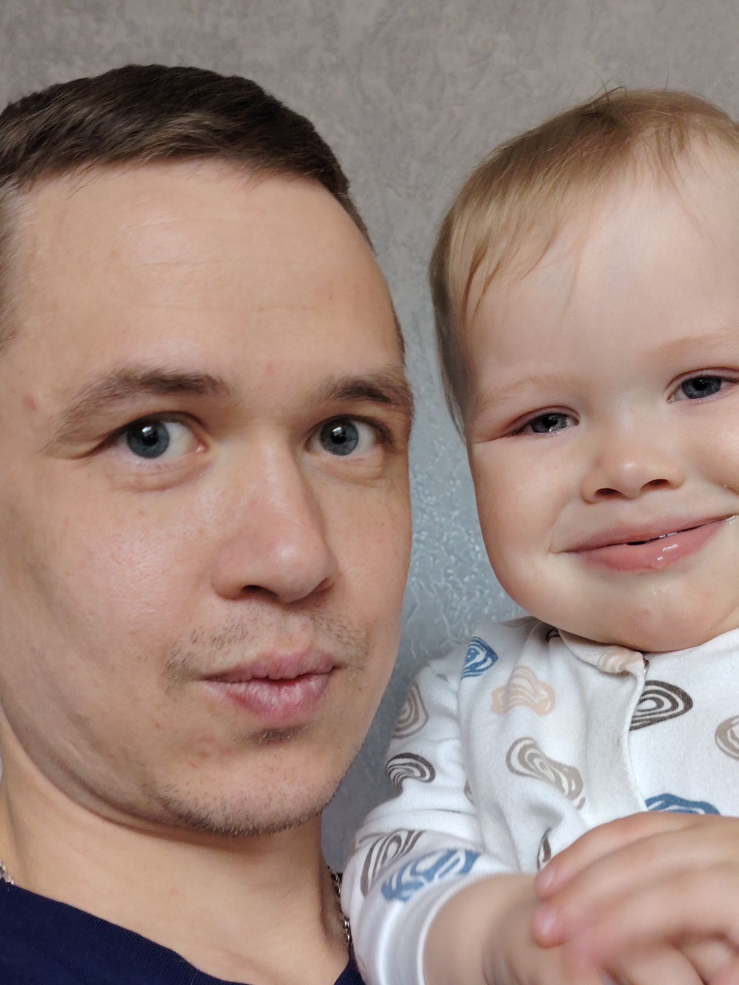 The image depicts a selfie of a man and a young child. The man is fair-skinned, with short brown hair, blue eyes, and a thin mustache. He wears a dark blue t-shirt and a silver chain. The child, with fair skin and blond hair, wears a white onesie with brown and blue patterns. The child appears to be drooling slightly. The background is a textured gray wall. The location is Chaikovsky, Russia.