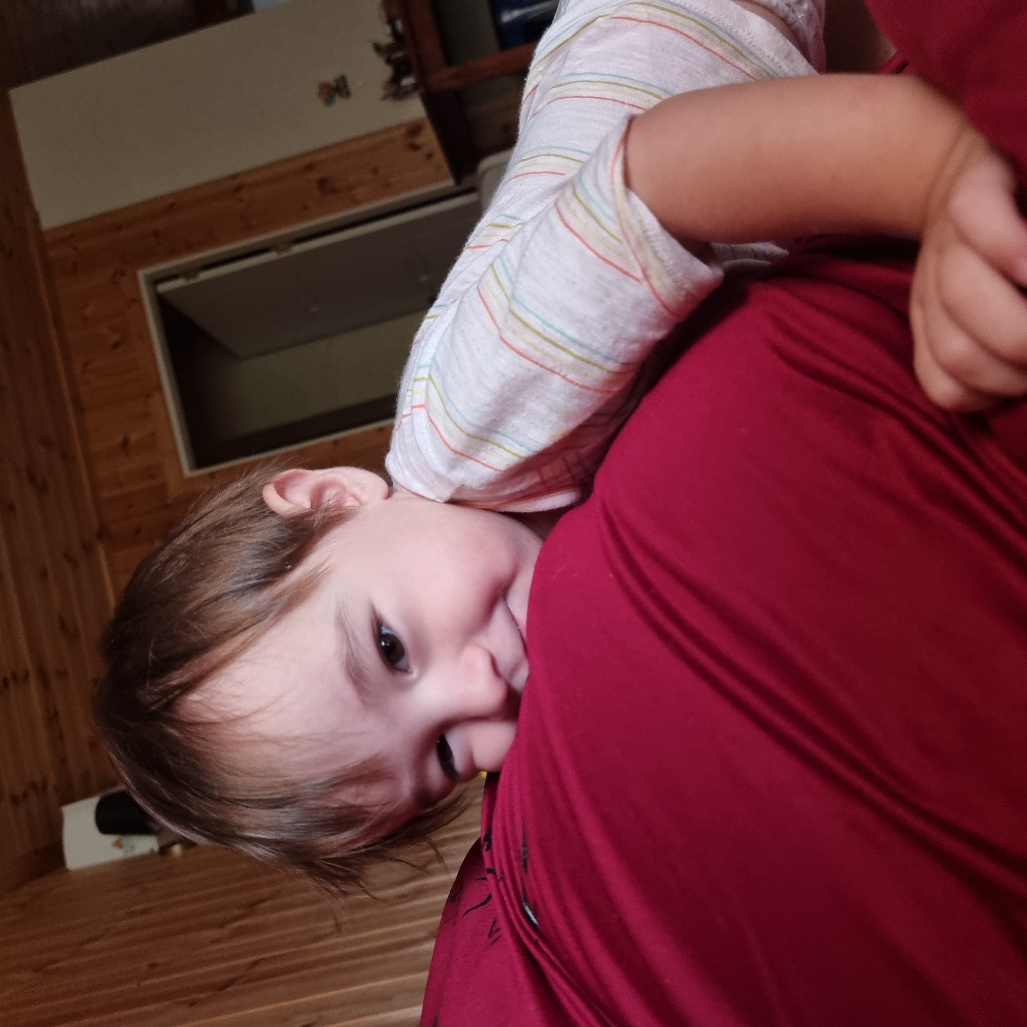 A close-up, eye-level shot shows a young child nestled into a large, deep red surface that appears to be a pillow or cushion. The child is positioned with their head resting on the red surface, looking directly at the camera with a playful expression. Their right arm is visible, resting on the red surface as well, with their hand lightly touching it. The child is wearing a light-colored t-shirt with thin, multi-colored horizontal stripes. The background is dimly lit and appears to be an interior room with wood-paneled walls and a doorway visible on the right. The overall impression is one of comfort and intimacy. The provided location of Dunnae-myeon, South Korea, suggests this scene is taking place in a home or similar domestic setting within that area.