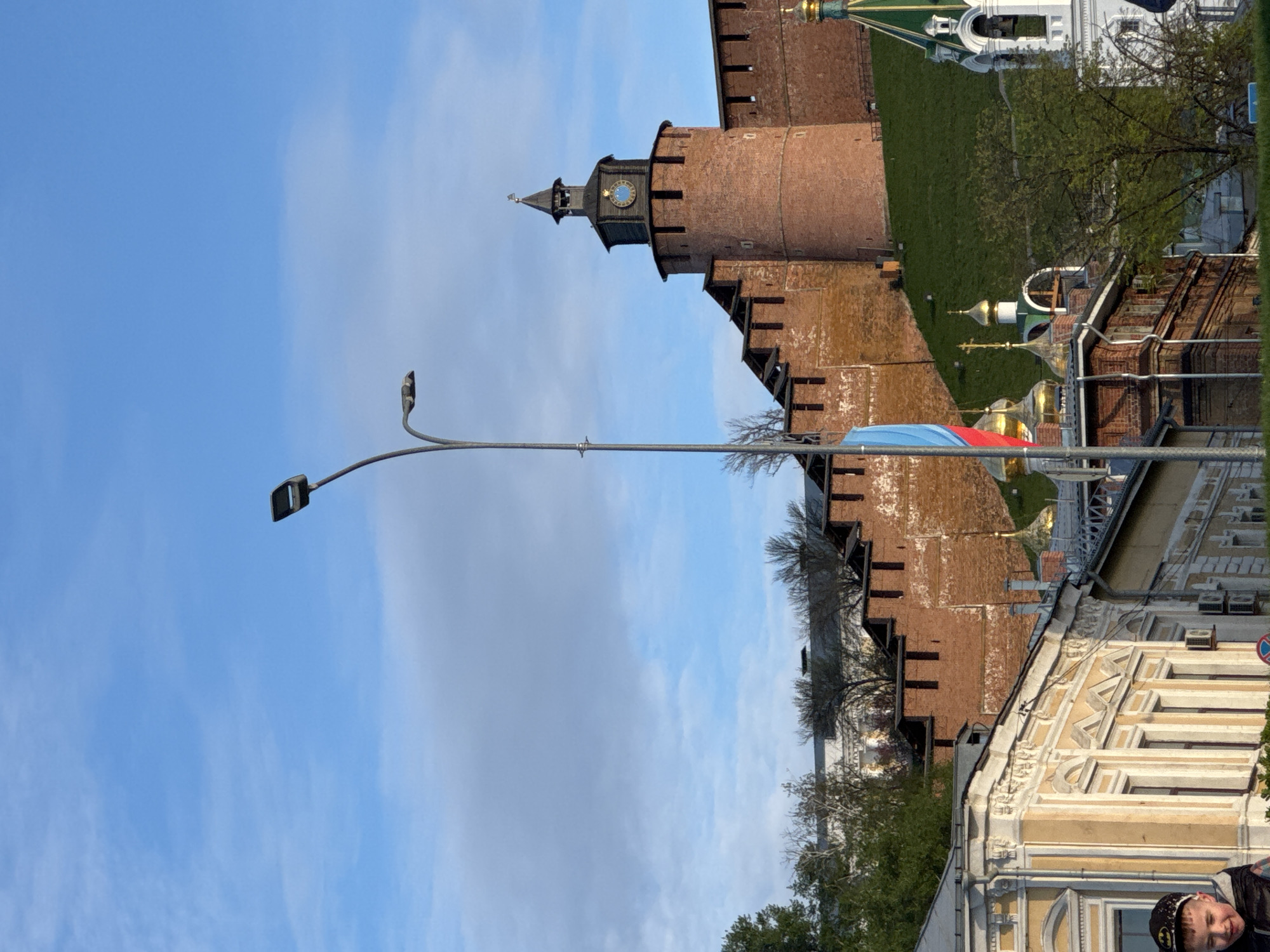The image shows a street scene in Nizhny Novgorod, Russia, dominated by the view of the Nizhny Novgorod Kremlin. The red brick walls and towers of the Kremlin are visible, including a tower with a clock face. A lamppost stands on the left of the image. Part of a building with decorative elements and windows is located in the lower right corner. The sky is blue with some clouds. A person's head is seen at the bottom right corner of the image.