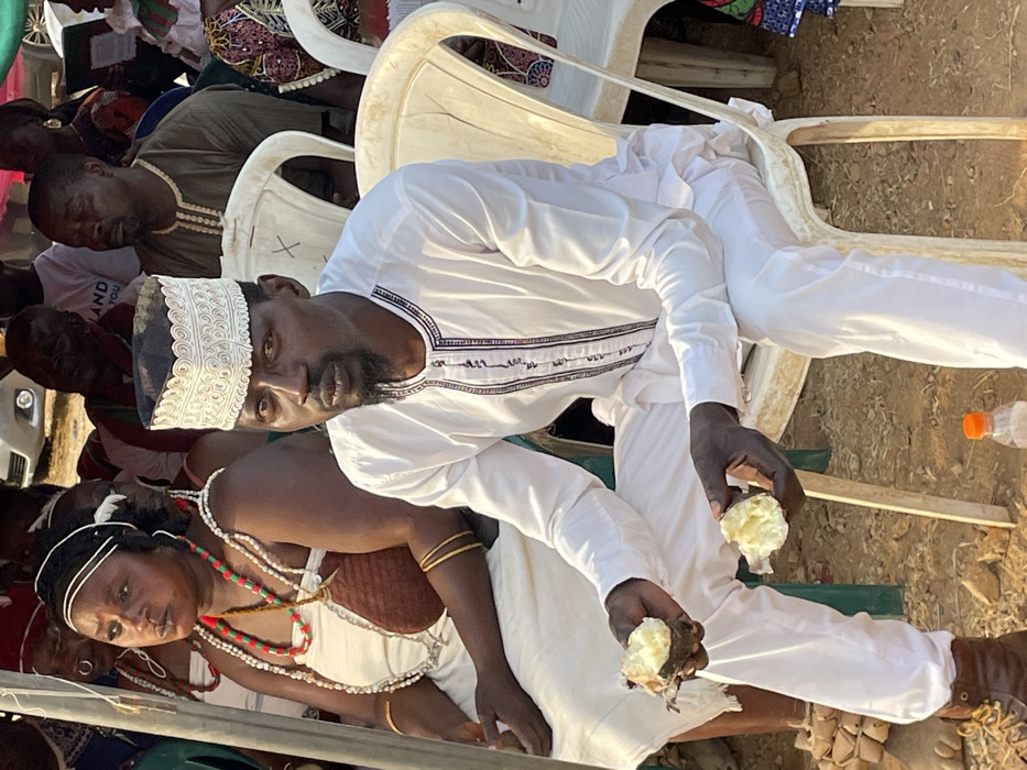 An outdoor scene captures a group of people gathered, likely for an event or social occasion, under what appears to be bright daylight. The ground is dry and earthy, typical of an outdoor setting.
In the foreground, a dark-skinned man is seated on a white plastic chair, looking directly towards the viewer. He wears a white, long-sleeved tunic with intricate dark embroidery around the neckline and chest, paired with white pants. A distinct white embroidered cap adorns his head. He has a dark beard and short dark hair. In both hands, he holds pieces of a light-colored, soft-looking food, possibly a cooked root vegetable like yam, suggesting he is eating or about to eat.
To the man's left (viewer's right, if the image were upright), a dark-skinned woman is also seated, her gaze directed downwards and slightly away from the camera. She is adorned in traditional attire, featuring a light-colored top with a brown, textured section across her chest, and multiple strands of vibrant bead necklaces in shades of red, green, white, and brown. Gold-colored bangles are visible on her left wrist.
Behind these two central figures, several other individuals are visible, mostly seated on similar white plastic chairs. One man directly behind the central man wears an olive green or khaki shirt and appears to be resting or looking downwards. Another person in the background wears a white t-shirt with partially visible text that seems to read "...AND YOU" or similar, though it's largely obscured.
A clear plastic water bottle with an orange cap is visible on the ground to the right side of the man, near his feet. The overall atmosphere appears casual and communal.