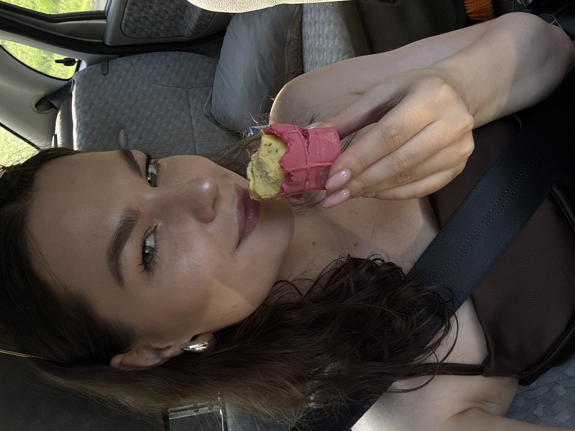 A young Caucasian woman with dark brown wavy hair is seated in the passenger seat of a car. She is wearing a dark brown camisole and a silver hoop earring in her left ear. The woman is holding a small ice cream cone in her right hand, which is positioned near her mouth. Her fingernails are painted a light pink color. The interior of the car is visible behind her, with grey patterned upholstery on the seats and a black seatbelt across her chest. The outdoor environment outside the car windows appears to be green and sunny, suggesting a daytime setting in a rural or suburban area. There is no specific indication of the location being Garevaya, Russia.