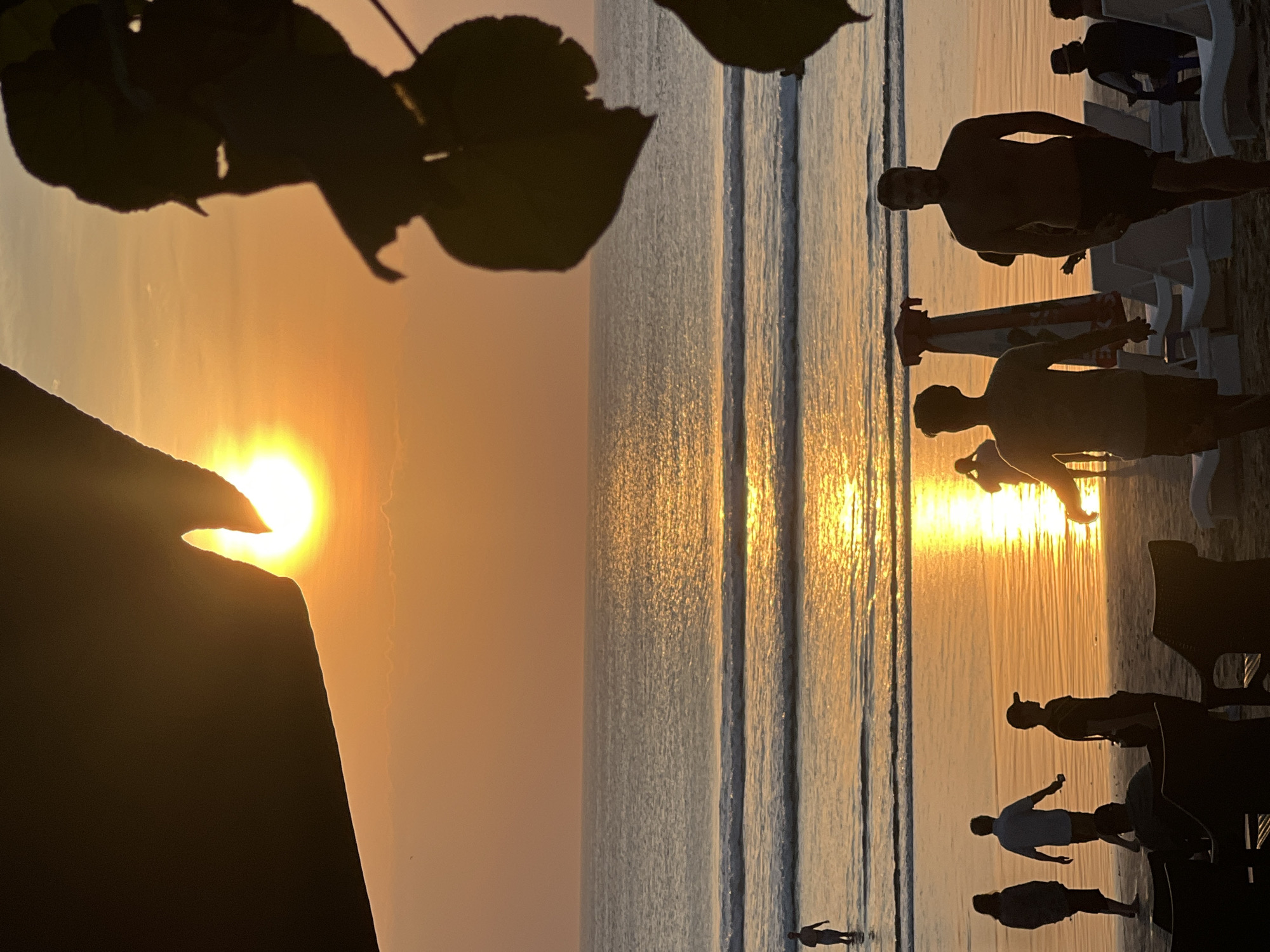 The image depicts a sunset on Kuta Beach, Indonesia, with the sun partially obscured by dark foliage in the foreground. The sky is a gradient of orange and yellow, reflecting on the calm ocean. Gentle waves break on the shore, creating a shimmering path of light. Several individuals are visible on the beach and in the shallow water, their figures silhouetted against the bright sunset. Some are walking, while others appear to be standing or wading. Beach chairs and umbrellas are scattered across the sand, suggesting a relaxed atmosphere. The overall impression is one of a serene and picturesque evening at a popular tourist destination.