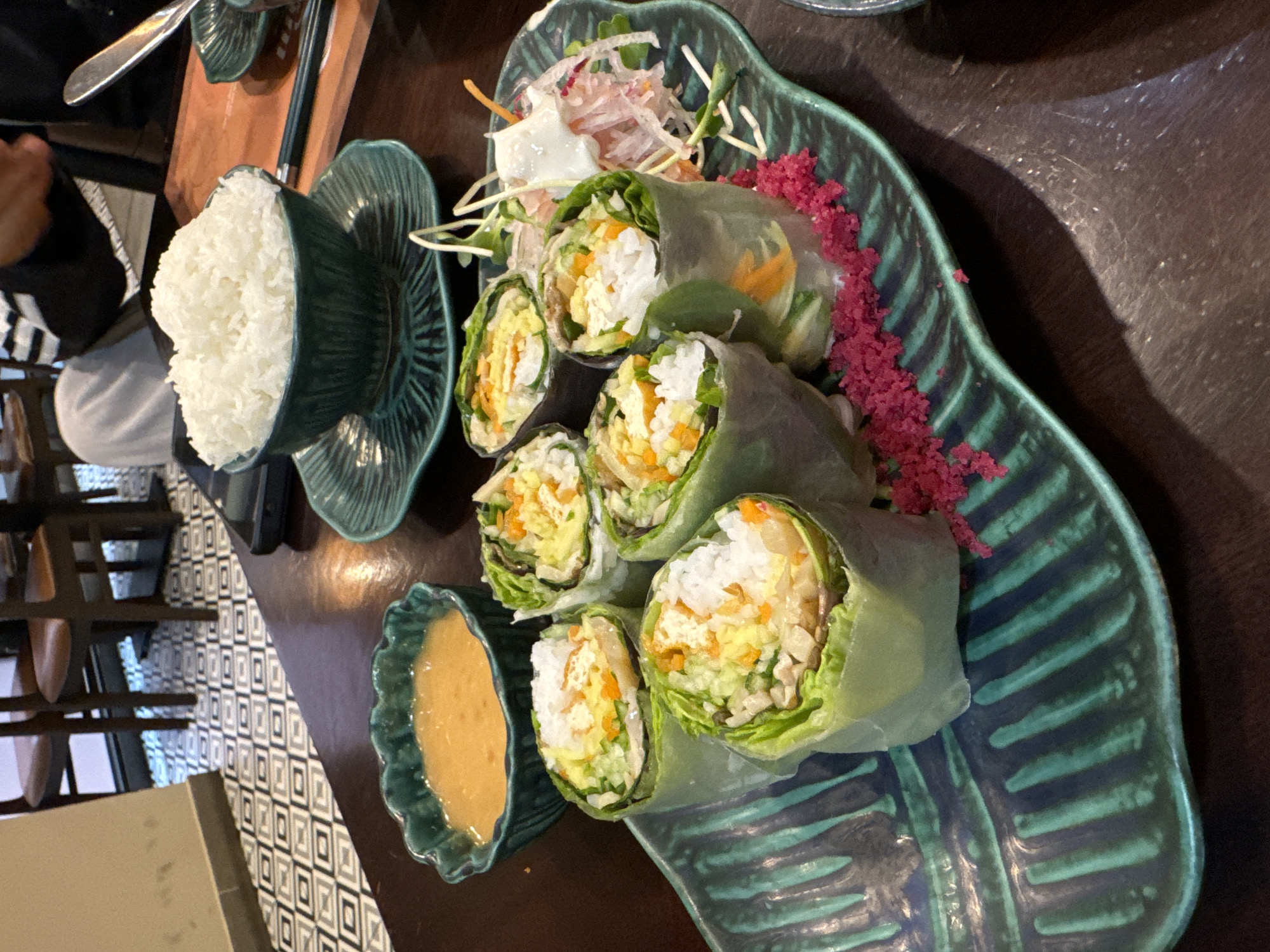 The image depicts a close-up overhead view of a table setting at a restaurant. The main subject is a platter of fresh spring rolls arranged on a dark green, leaf-shaped plate with ribbed detailing. The spring rolls are translucent, revealing their fillings of rice, shredded vegetables, and possibly other ingredients. To the left of the platter is a small, decorative green bowl filled with a light orange dipping sauce. In the background, a larger dark green bowl filled with white rice is visible, resting on a matching saucer. Additional tableware, including a spoon and chopsticks, are also present on the dark wooden table. The flooring in the background features a geometric pattern of black, white, and grey squares. The lighting suggests an indoor setting, possibly during the daytime. The scene is likely located in Phan Thiết, Vietnam, a city known for its coastal beauty and culinary offerings.