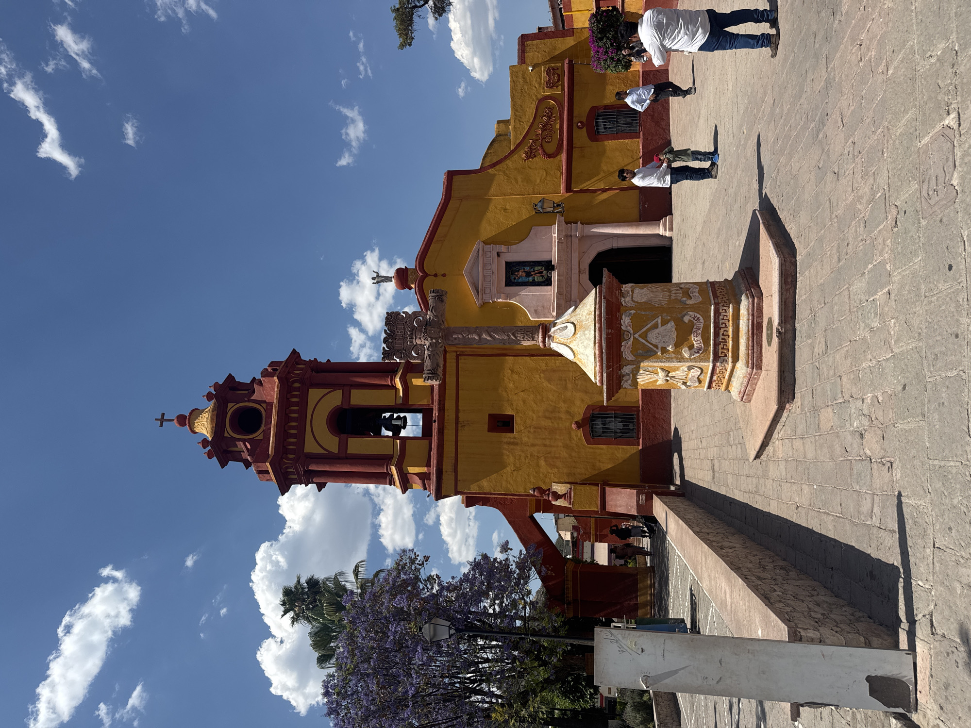 The image depicts a colorful church in Bernal, Mexico. The church is primarily yellow with red accents on the tower and trim. In front of the church stands a stone cross monument. Several people are visible near the entrance of the church. The sky is blue with scattered white clouds. A tree with purple flowers is visible at the bottom left corner of the image. The ground is paved with stone.