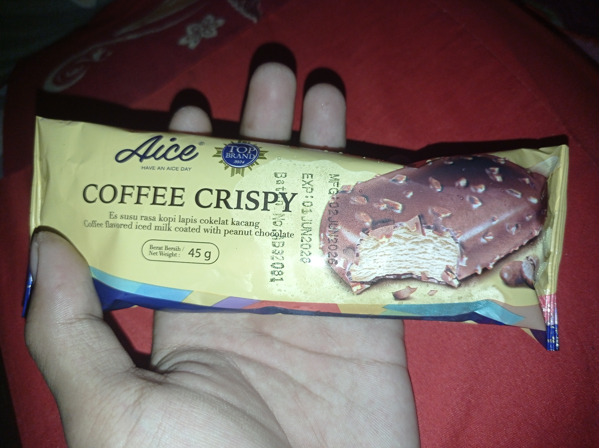 A close-up, slightly blurry, eye-level shot shows a hand holding a wrapper for an "Aice Coffee Crispy" ice cream bar. The wrapper is predominantly yellow with blue and purple accents at the bottom. The ice cream bar itself, partially visible, is chocolate-coated with what appear to be nuts. Text on the wrapper includes "Aice," "HAVE AN AICE DAY," "COFFEE CRISPY," "Es susu rasa kopi lapis cokelat kacang," and "Coffee flavored iced milk coated with peanut chocolate." Nutritional information such as "Berat Bersih / Net Weight : 45 g" is also present. Additional text indicating production and expiration dates (MFG: 02.JUN.2026, EXP: 02.JUN.2028) is visible. The background is a textured red surface, possibly a tablecloth or blanket. The context of Sumbernadi, Indonesia, is not directly evident from the image.