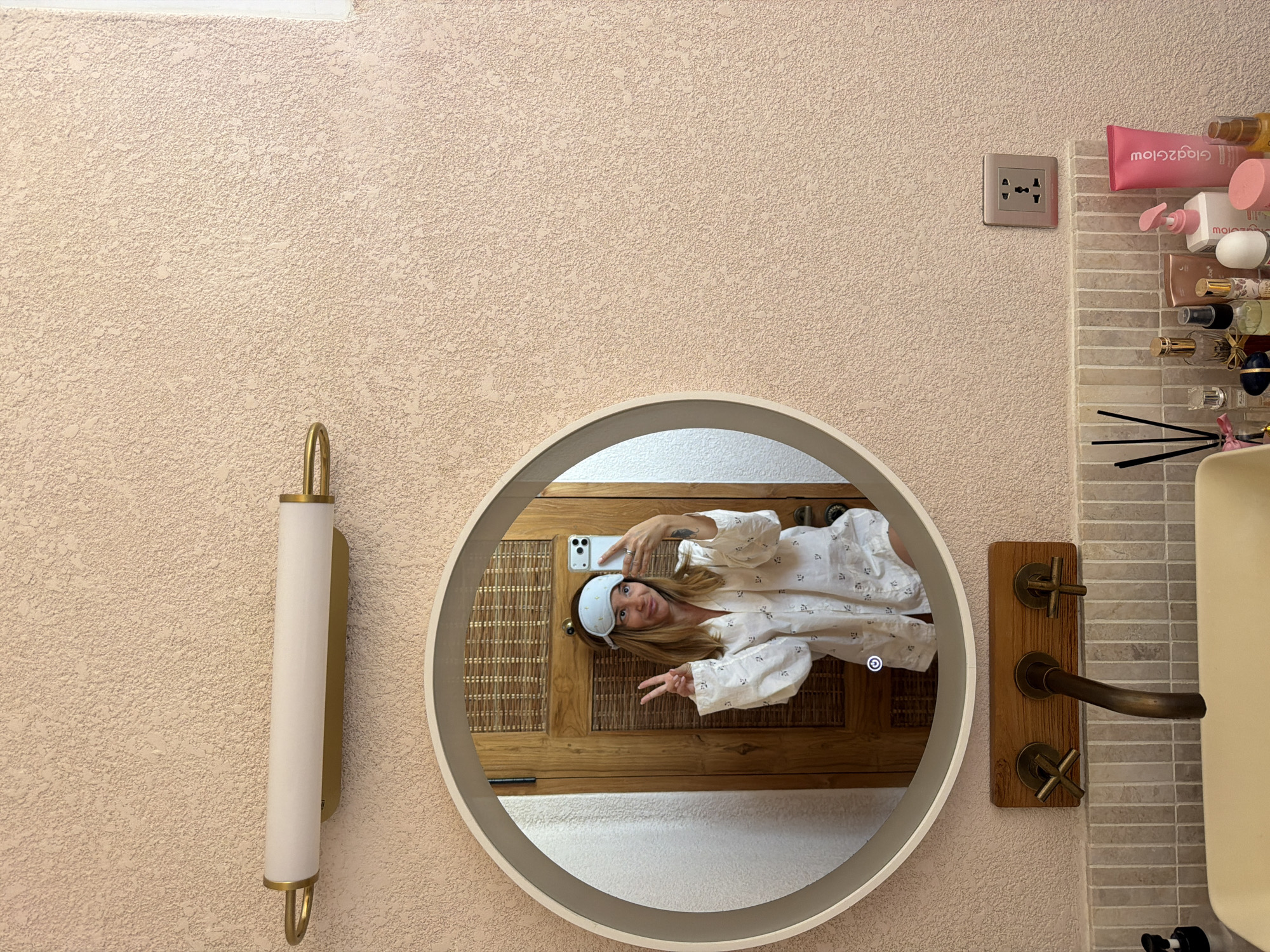 In a bathroom with textured pale pink walls, a woman is taking a mirror selfie. She is wearing a light blue sleep mask and a white long-sleeved pajama top with a small dark pattern. She is holding up two fingers in a peace sign and smiling. A round mirror with a thin, light-colored frame is visible, as is a gold and white light fixture above it. Below the mirror, there is a wooden panel with a bronze faucet and two cross-shaped handles. A sink is partially visible. On the counter to the right, there are various cosmetic products including a pink tube labeled "Glad2Glow," a pink pump bottle, and several other bottles and containers. A multi-socket electrical outlet is mounted on the wall to the right of the cosmetics. The overall setting suggests a domestic bathroom.