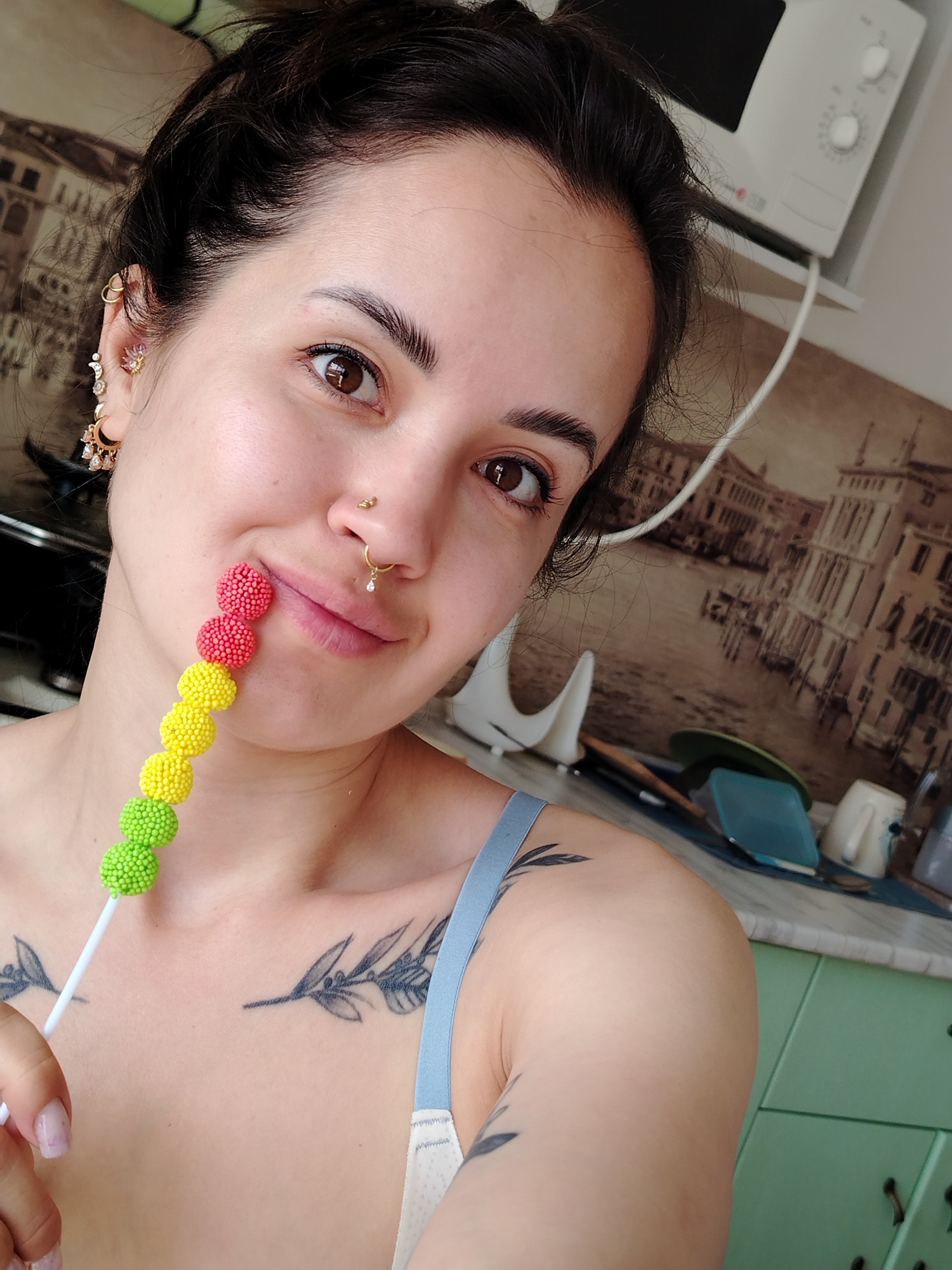 A young woman takes a selfie in what appears to be a kitchen in Noviy, Russia. She has dark hair, dark brown eyes, and light skin, looking directly at the camera with a gentle, inviting smile. Her hair is casually tied up, and she wears multiple piercings, including ornate gold earrings with moon and star motifs, a nose stud, and a septum ring with a dangling charm. Delicate leaf or branch tattoos are visible on her chest and shoulder. She is holding a white stick adorned with a colorful string of textured, spherical candies (red, yellow, and green), with one red candy resting playfully against her upper lip.
The background shows a domestic kitchen environment, illuminated by natural daylight. A white microwave is visible on a shelf to the upper right. A prominent sepia-toned backsplash wallpaper behind her depicts an elaborate cityscape, possibly Venice. On the countertop below, various kitchen items such as dishes, a white mug, and a silver kettle are discernible, alongside green kitchen cabinets at the bottom right of the frame. No text is visible in the image.
