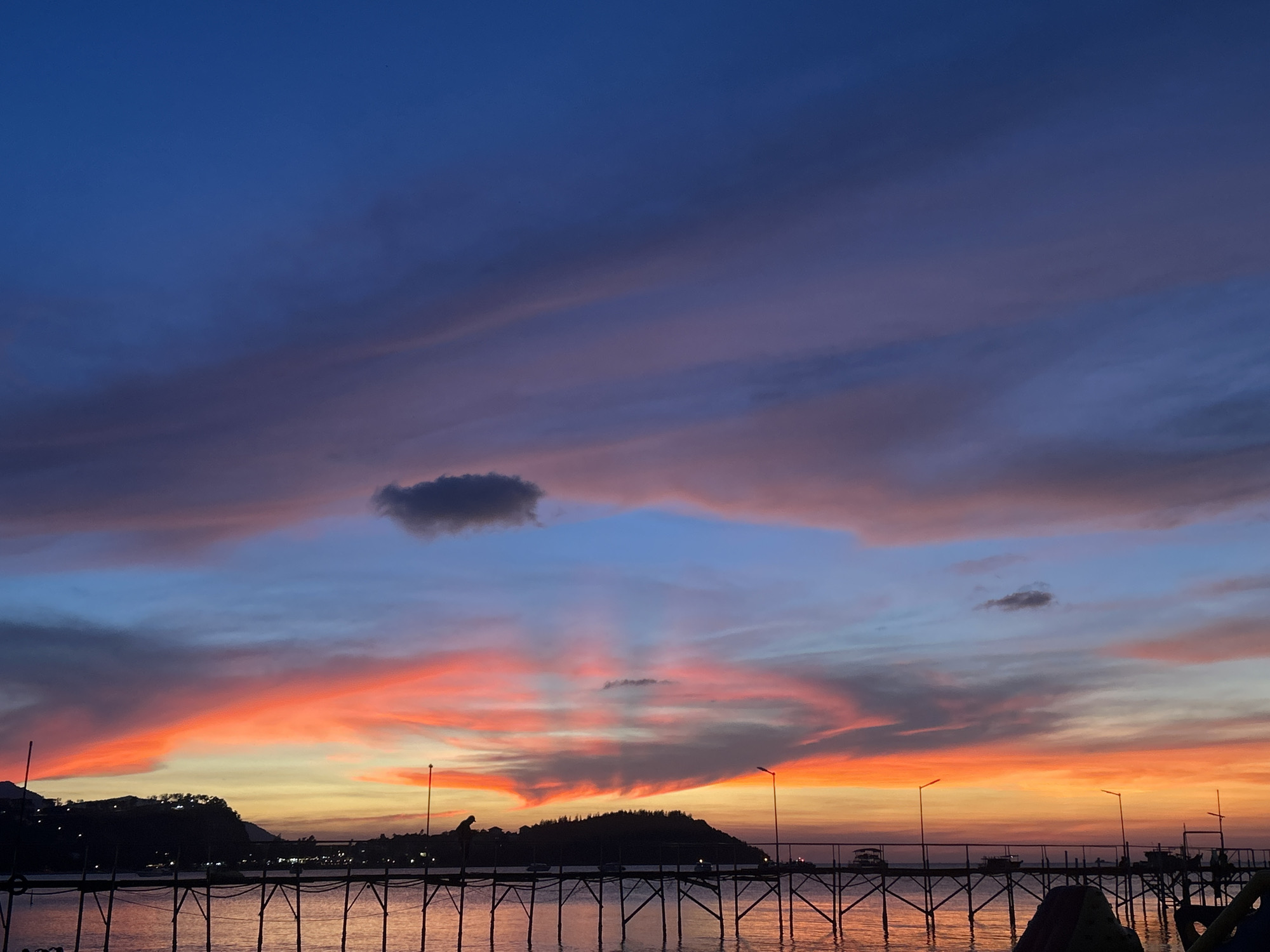 Captured in Baan Bang Rak, Thailand, the photograph showcases a dramatic sunset over the water. The sky transitions from a deep blue at the top to hues of purple, pink, orange, and yellow closer to the horizon. Clouds are streaked across the sky, with one small, isolated cloud above the horizon. A dark landmass is visible in the middle ground, and a pier extends into the water. Streetlights are spaced along the pier, and their reflections shimmer on the water's surface. There are hints of human presence on the pier towards the right side of the frame. The overall scene captures the tranquil beauty of dusk.