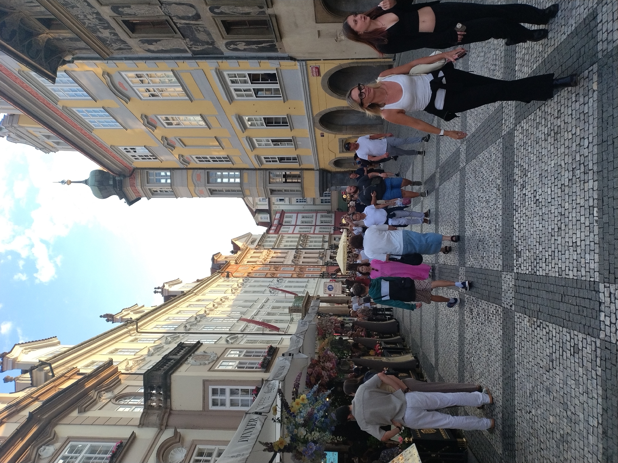 The image shows a street scene in Prague, Czechia, likely during the day. The architecture suggests an older European city with buildings featuring ornate facades, balconies, and distinct window styles. The street is paved with cobblestones and lined with shops and restaurants, some of which have outdoor seating and awnings decorated with flowers. Several people are walking along the street, some in groups and some individually. They are dressed in casual summer clothing. In the foreground, a man in a light-colored shirt and white pants is standing with his hands on his hips, facing away from the camera. To his right, a woman in a white tank top and black pants is walking towards the camera, wearing glasses and a necklace. To her right, another woman in a black top and black pants is walking, looking towards the camera. The sky is visible with some clouds, and the sunlight casts shadows on the buildings and the street.