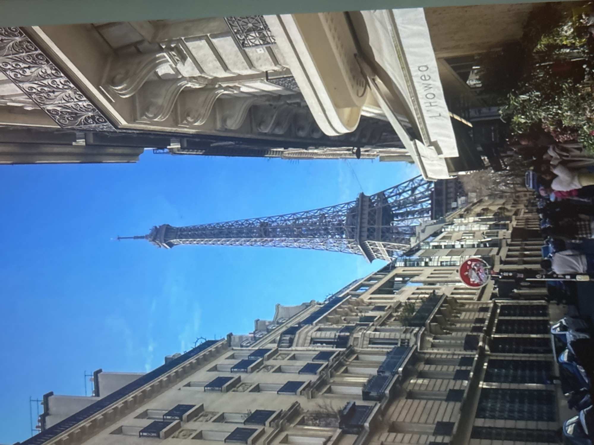 The image shows a street in Paris, France, with the Eiffel Tower visible in the background. The street is lined with buildings with ornate balconies and windows. There are people walking on the sidewalk and sitting at outdoor cafes. Cars are parked along the street. The Eiffel Tower is a tall, iron lattice tower located on the Champ de Mars in Paris. It is one of the most recognizable landmarks in the world. The image was taken on a sunny day, with a clear blue sky.
