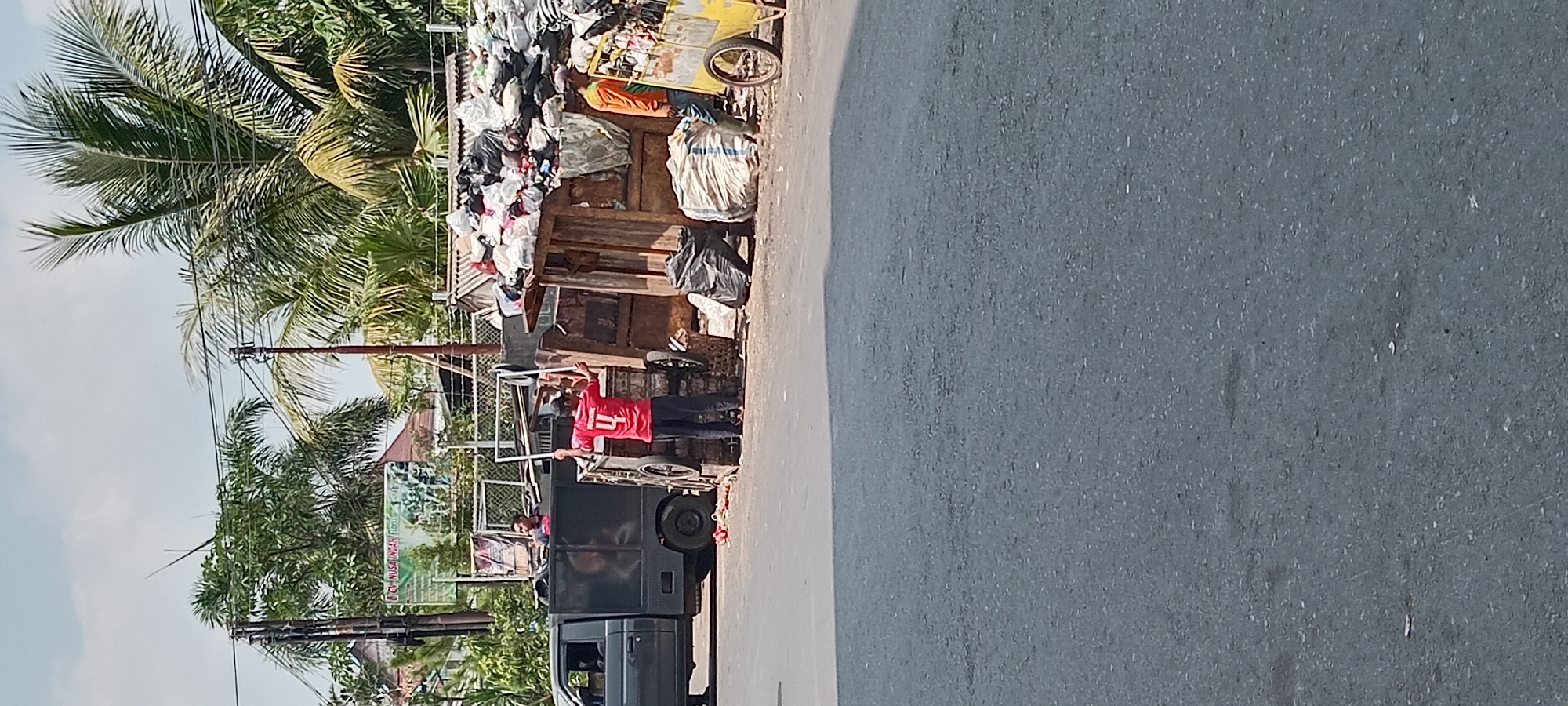 A man in a red shirt with the number 4 on the back stands by a wooden structure overflowing with trash bags. He is holding a white frame. To his left, a dark grey pickup truck is parked. Another man in an orange shirt stands near a yellow cart loaded with trash. The scene appears to be a roadside refuse collection point in Tasikmalaya, Indonesia, with palm trees and power lines visible in the background. The ground is paved, and the sky is overcast.