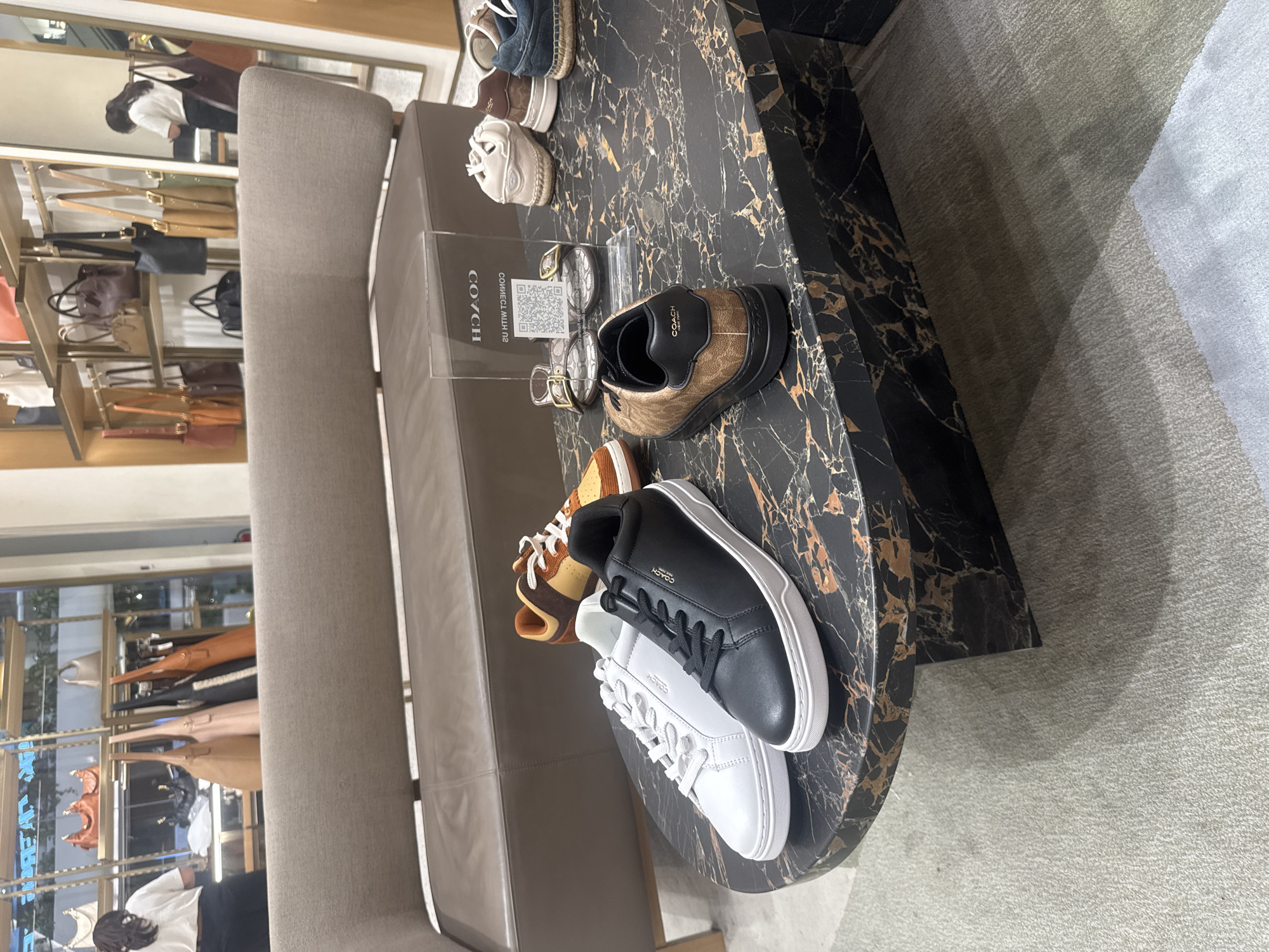 This image shows a retail display of Coach footwear in a store. The foreground features a selection of sneakers on a black marble table. There are three pairs of sneakers: a brown and gold patterned pair, a black pair with a white sole, and a white pair with a white sole. All three pairs have white laces and the Coach logo visible. The black marble table has a modern, luxurious feel.
Behind the table, a grey upholstered bench is visible. In the background, shelves are stocked with various Coach handbags in different colors and styles, including tan, pink, and black. A person is partially visible in the reflection of a mirrored surface to the left of the image, appearing to be a female wearing a white top and dark bottoms.
The overall setting is a well-lit retail environment, likely a Coach boutique or department store section. The arrangement suggests a product display designed to attract customers. The time of day is not explicitly indicated, but the bright lighting suggests it is daytime. The scene appears to be indoors, in a climate-controlled setting. The visible text includes the "COACH" logo on the shoes and on a small sign placed on the marble table, which also has a QR code printed on it. The location is identified as Denpasar, Indonesia.