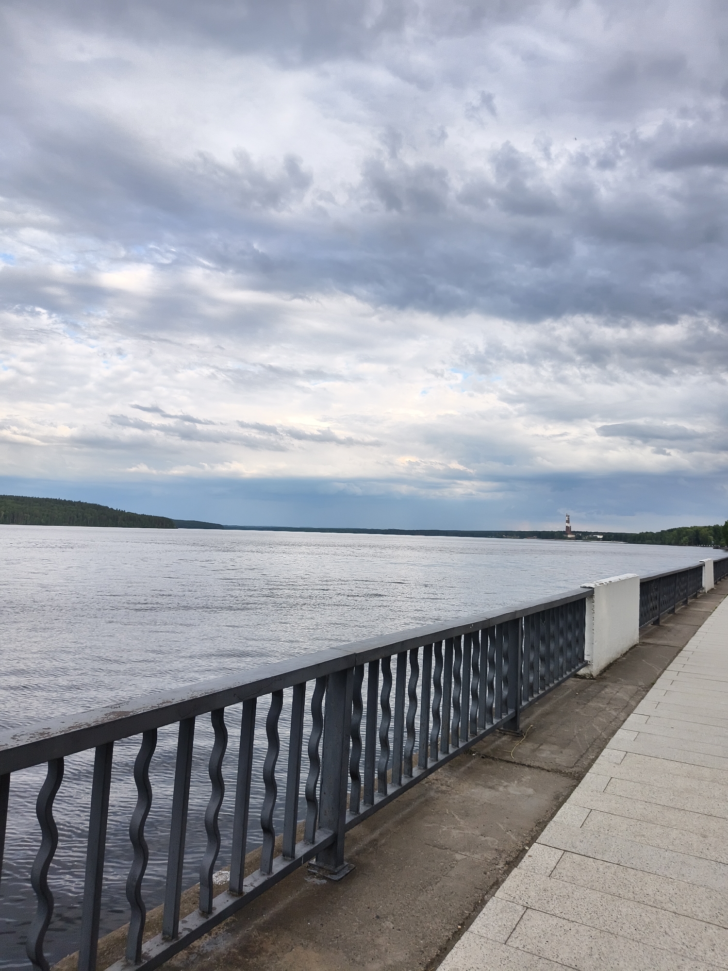 This media file is a landscape photograph taken from a waterfront promenade. The foreground features a dark metal railing with a wavy, decorative pattern, separating the promenade from a wide body of water. The promenade itself is paved with light-colored, rectangular tiles. Beyond the railing, the calm, grey water stretches towards a distant, tree-covered shoreline on the left. On the right, the water recedes into the distance, and a lighthouse with red and white bands is visible in the far distance.
The sky is heavily clouded with shades of grey and white, suggesting an overcast or impending stormy weather. The overall mood is somber and peaceful, with the vastness of the sky and water dominating the scene. The time of day appears to be late afternoon or early evening, given the subdued lighting. The location cues suggest a riverside or lakeside promenade in Chaikovsky, Russia, with the lighthouse being a notable landmark. There is no visible text in the image.