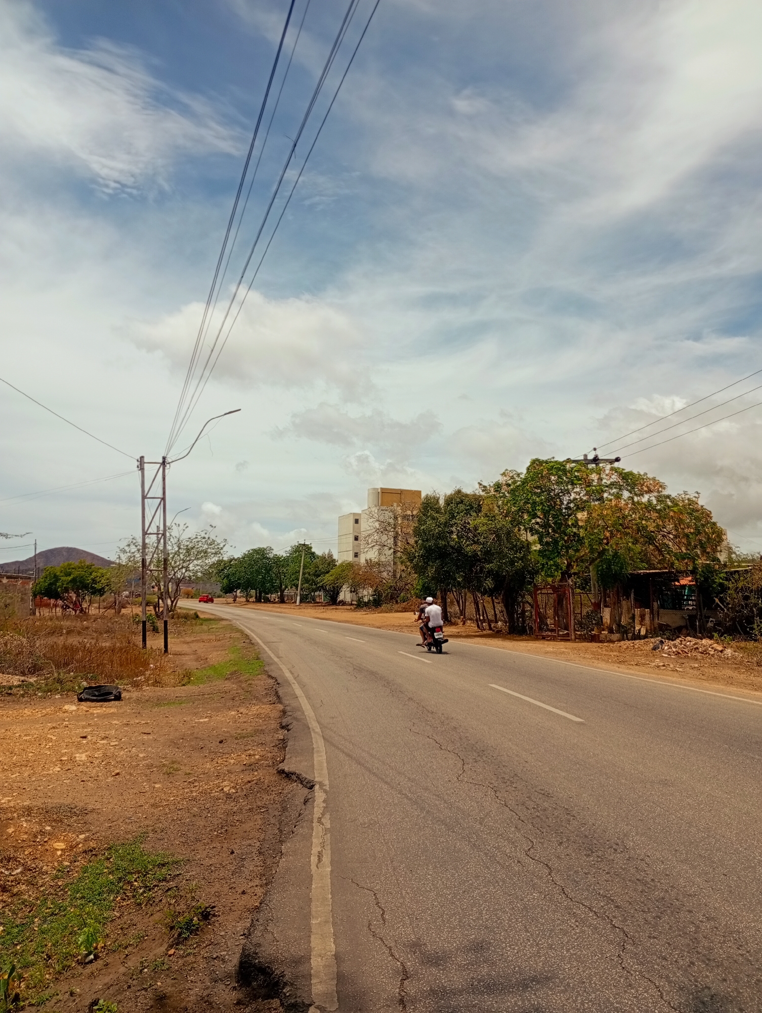 The image depicts an outdoor scene in Altagracia, Venezuela, featuring a curving asphalt road under a partly cloudy sky. A single individual wearing a white t-shirt and a white helmet is observed riding a dark-colored motorcycle away from the viewer in the right lane of the road. The road surface exhibits multiple cracks. A white solid line runs along the right edge of the road, and a broken white line is visible near the center.
To the left of the road, a tall metal utility pole with a street light extends upwards, supporting multiple overhead power lines that traverse the upper portion of the frame diagonally from left to right. Below the pole, the unpaved shoulder consists of reddish-brown earth with sparse green vegetation, and a dark, possibly fabric-like, object rests on the ground. A red car is partially visible in the distance on the left side of the road.
On the right side of the road, a dense line of trees with varying foliage shades lines the shoulder. Behind these trees, a multi-story white building with yellow upper sections is visible in the mid-ground. Further right and partially obscured by trees, a smaller, dilapidated structure with a corrugated roof is present. Another utility pole stands on the right side of the road further down. The sky features scattered white cumulus clouds against a blue background. The overall environment appears semi-urban or rural, characterized by dry, earthy terrain on the road shoulders and a mix of natural vegetation and human infrastructure.