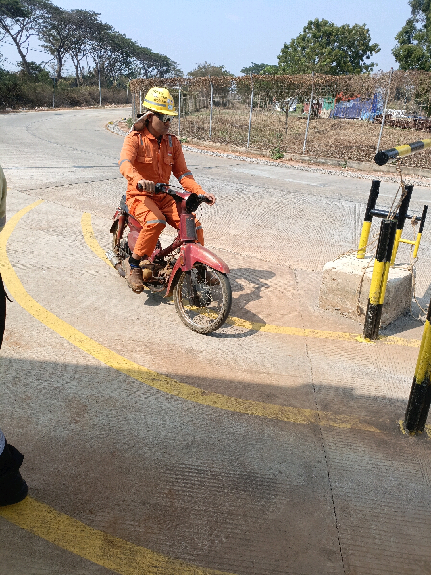 In Jenu, Indonesia, a person wearing an orange jumpsuit and a yellow hard hat is riding a red motorcycle on a paved road marked with yellow lines. The person is facing forward, with their hands on the handlebars. The road curves to the left, and a chain-link fence with some vegetation behind it is visible in the background. There are also some bollards with yellow and black stripes on the right side of the image. The sky is clear and blue.