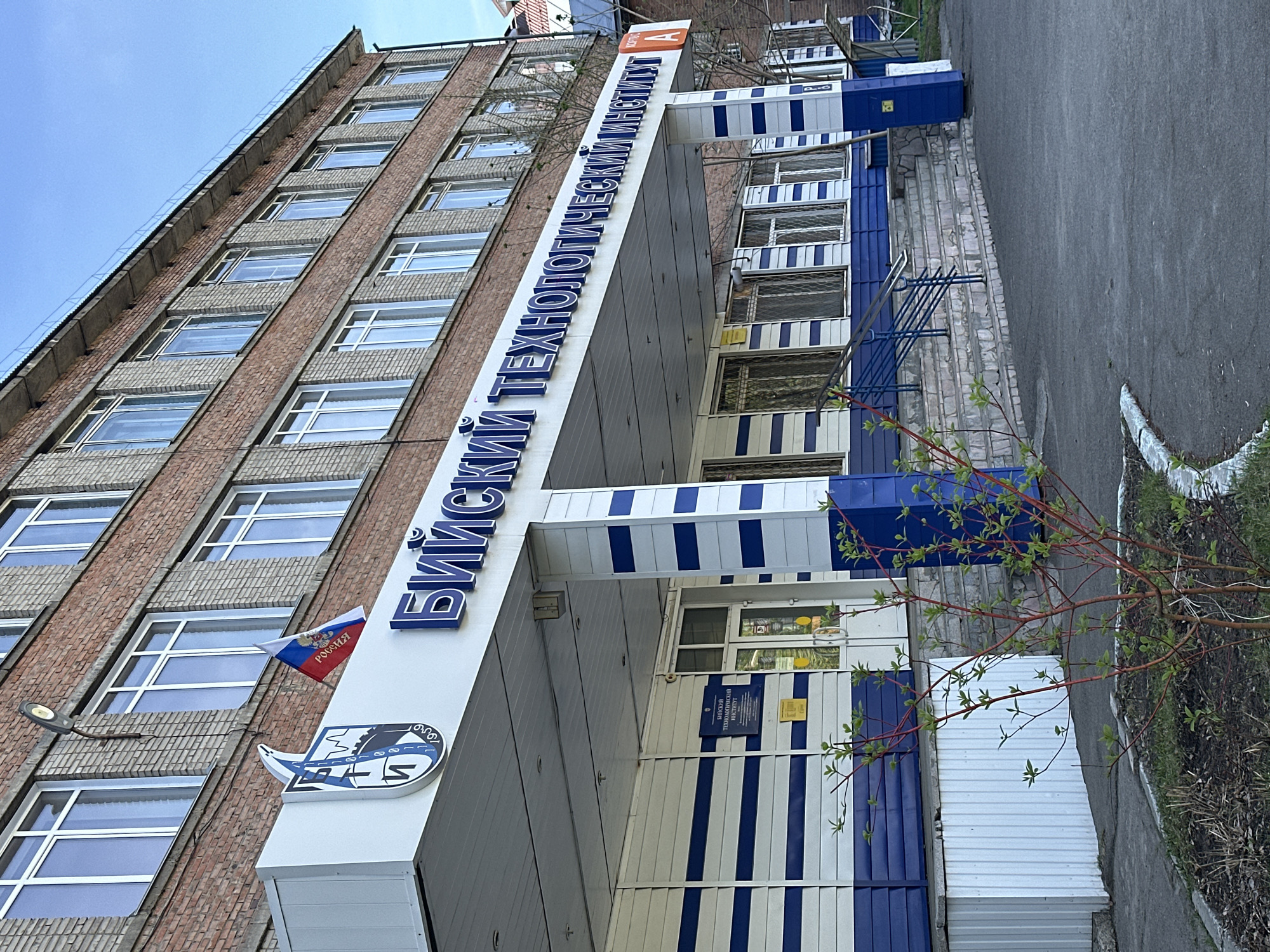 The image captures the exterior of a multi-story brick building, likely an educational or technological institute, under a clear blue sky during daylight hours. The building facade features a prominent entrance area adorned with blue and white striped paneling.
The main subject is the building itself, constructed primarily of reddish-brown bricks with rows of large, rectangular windows set in light-colored frames. The lower section around the entrance is covered with white and blue panels, including horizontal blue stripes on a white background, creating a distinctive pattern. Above the entrance, a large white sign with raised blue Cyrillic letters proudly displays "БИЙСКИЙ ТЕХНОЛОГИЧЕСКИЙ ИНСТИТУТ" (Biysk Technological Institute). A small orange 'A' logo is visible on the upper right of this sign.
To the left of the main entrance, a Russian flag is mounted on the brick wall, clearly displaying the word "РОССИЯ" (Russia) on its white stripe. Below the flag, there's a circular emblem or logo with blue and white elements, featuring what appears to be a stylized industrial or technological graphic. A small, dark blue sign is also affixed near the entrance, repeating the name "БИЙСКИЙ ТЕХНОЛОГИЧЕСКИЙ ИНСТИТУТ."
The immediate surroundings include a paved asphalt area to the right, suggesting a driveway or parking space, and some sparse greenery with new shoots in the foreground, indicating early spring or summer. No people are visible, and no specific activity is taking place; the scene presents a static view of the institute's entrance. The overall impression is one of a functional, institutional building in Biysk, Russia, as indicated by the text and flag.