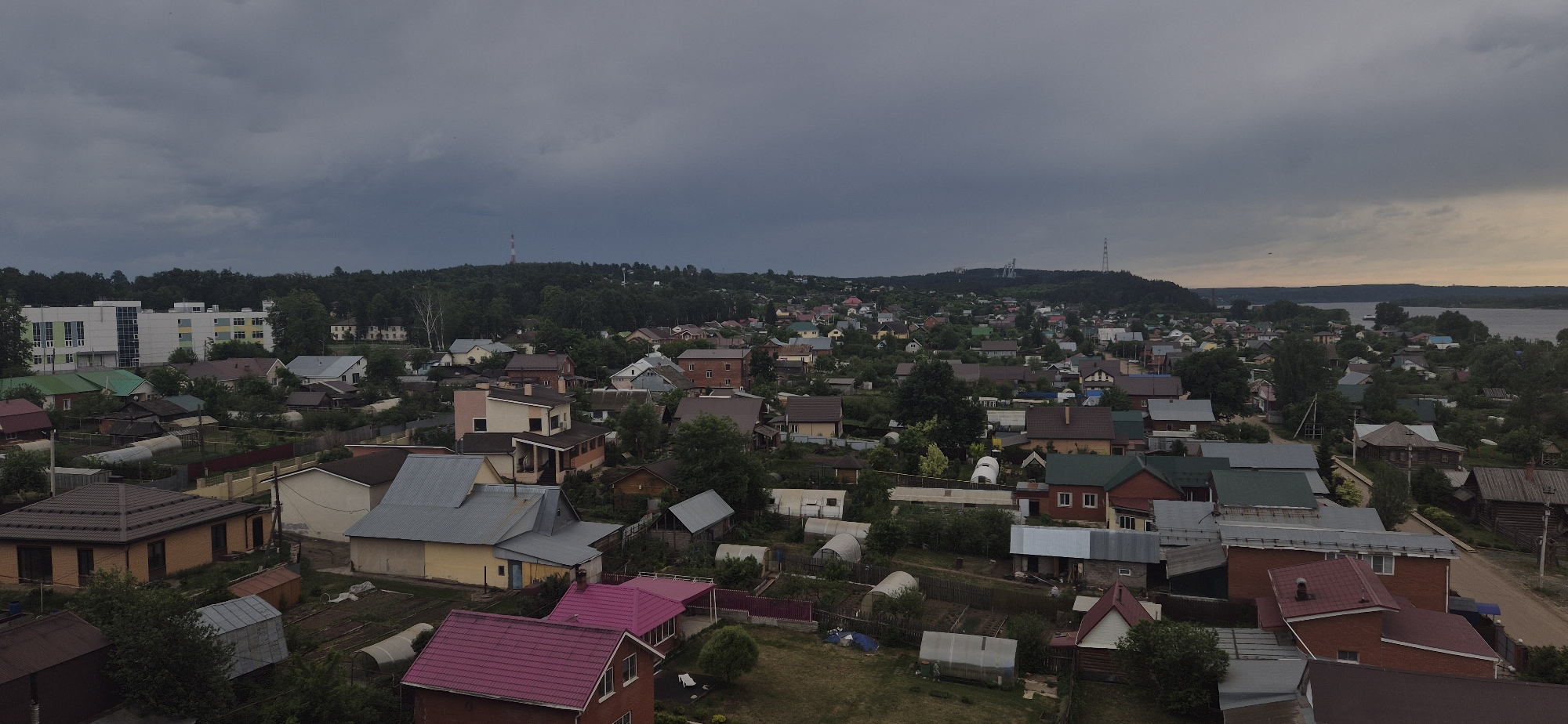 The image shows a general view of the Russian city of Chaikovsky, Perm Krai, with numerous residential houses surrounded by lush vegetation under an overcast sky. In the distance, there is a large river or lake, and on the horizon, towers and structures can be seen on a hill. The roofs of the houses are varied, with different colors, including pink. In the foreground, gardens and greenhouses are visible, indicating an agricultural presence within the residential area. The overall scene suggests a suburban or rural setting. The lighting indicates that it may be late afternoon or early evening.